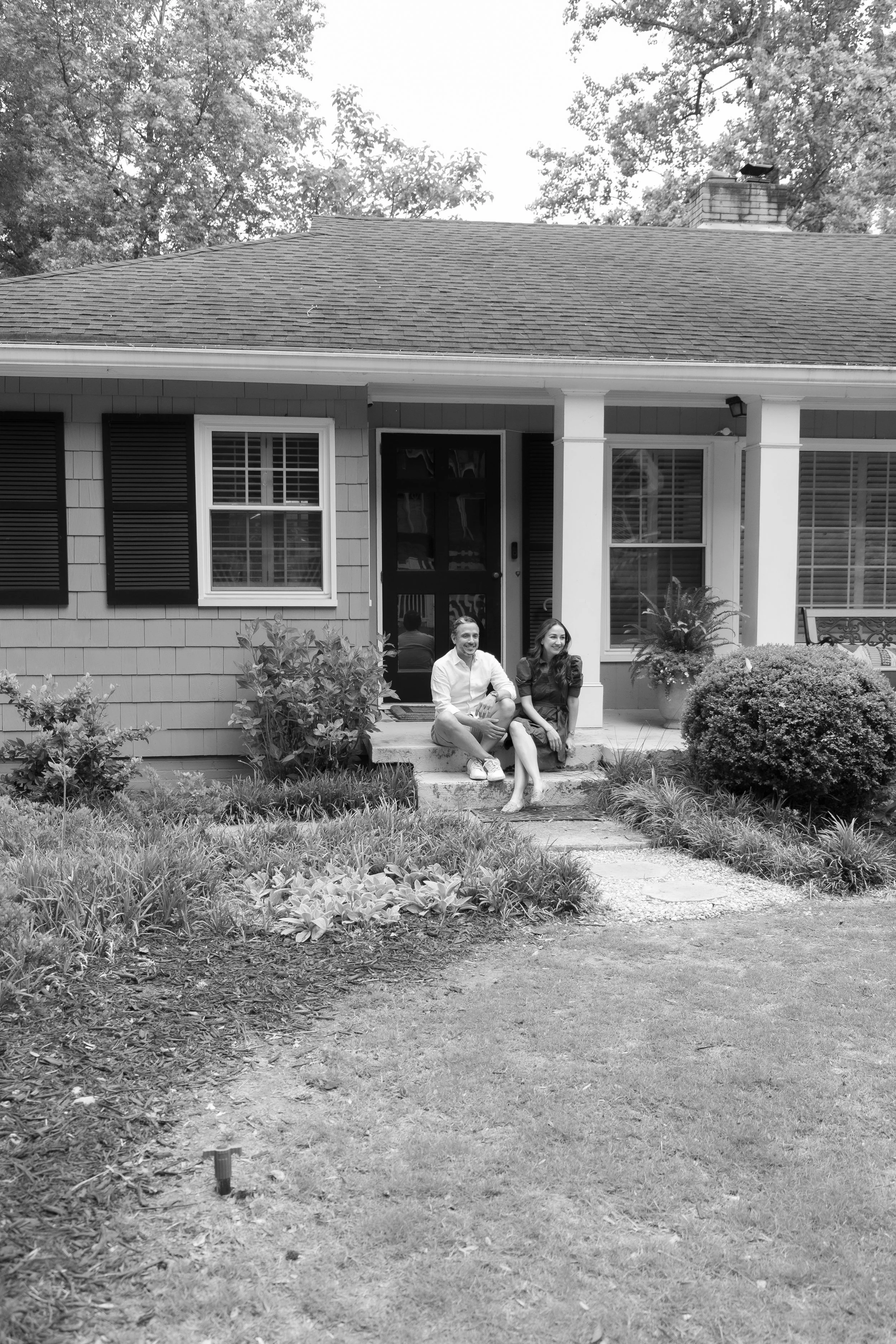 A black and white photo of two people sitting on front steps of a house, smiling. The house has a porch, window shutters, and a garden in front with plants and shrubbery. The yard has a dirt pathway leading up to the steps.