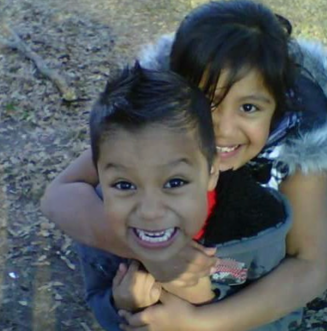 Two children playing outdoors, smiling and hugging each other, with fallen leaves and dirt ground in the background.