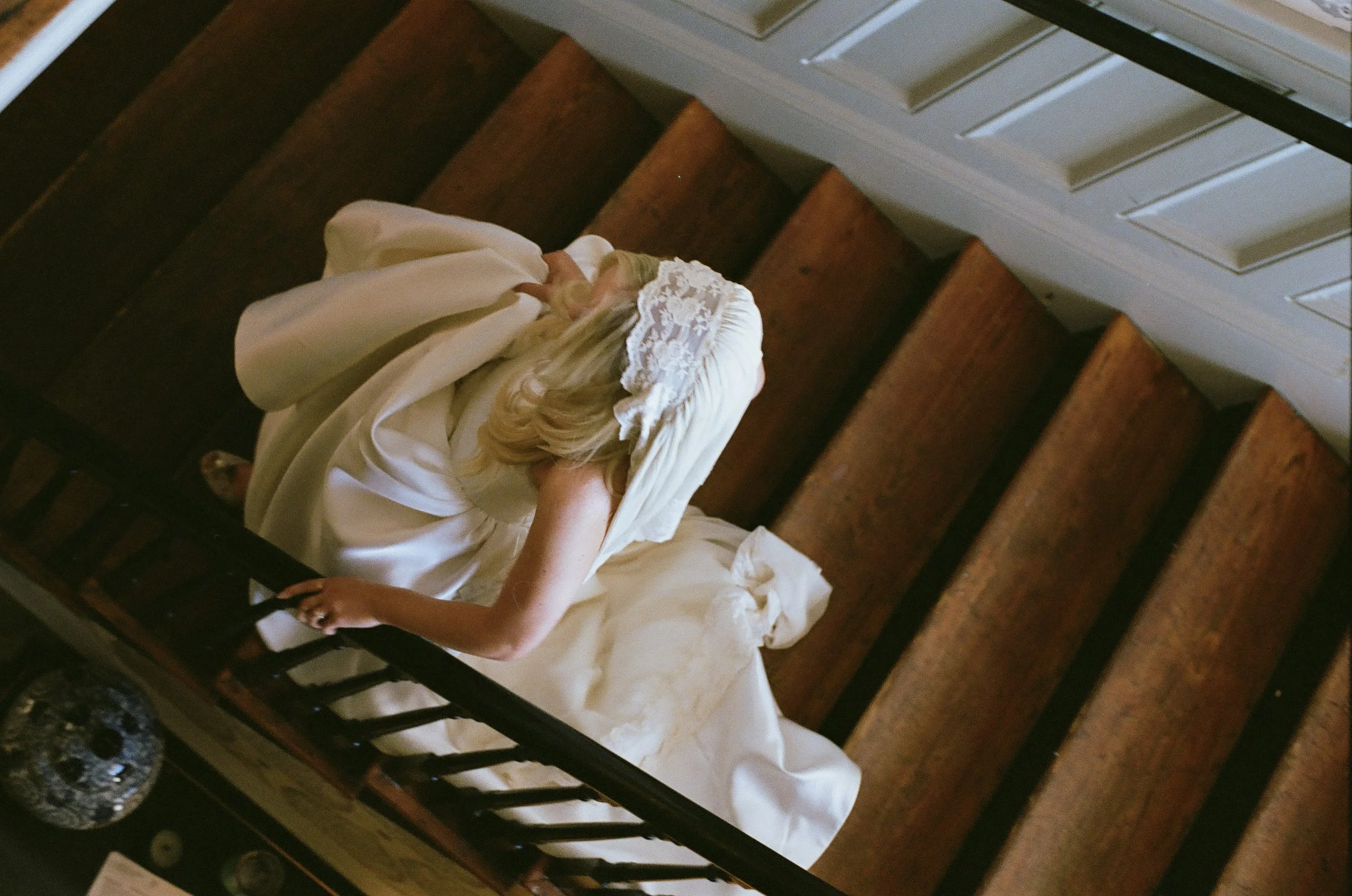 A woman with long blonde hair wearing a vintage dress and lace bonnet walking down wooden stairs in a historic house or museum.