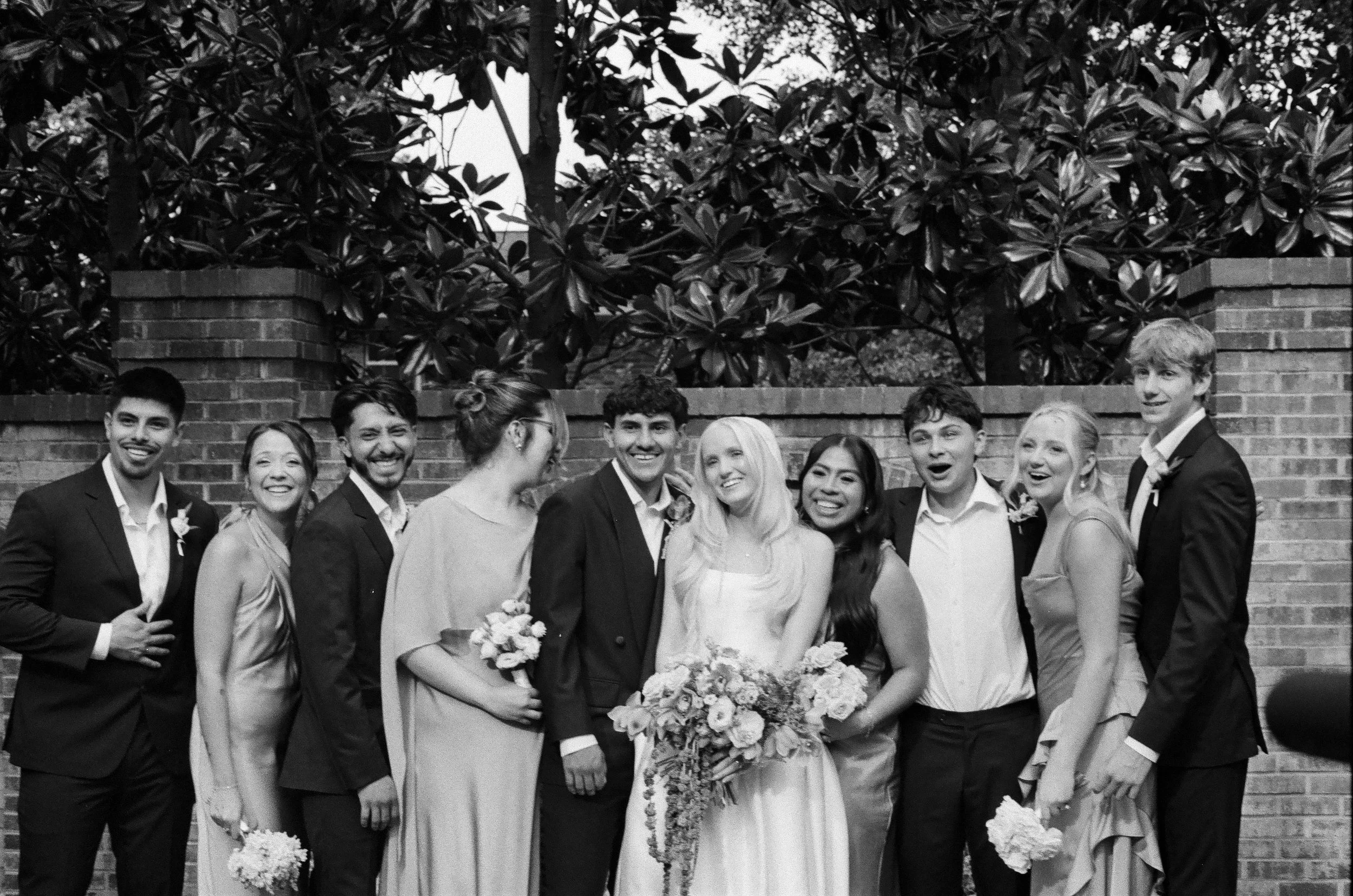 A group of people at a wedding, standing outdoors against a brick wall and leafy trees. The bride and groom are in the center, smiling and holding flowers, surrounded by friends in formal attire.