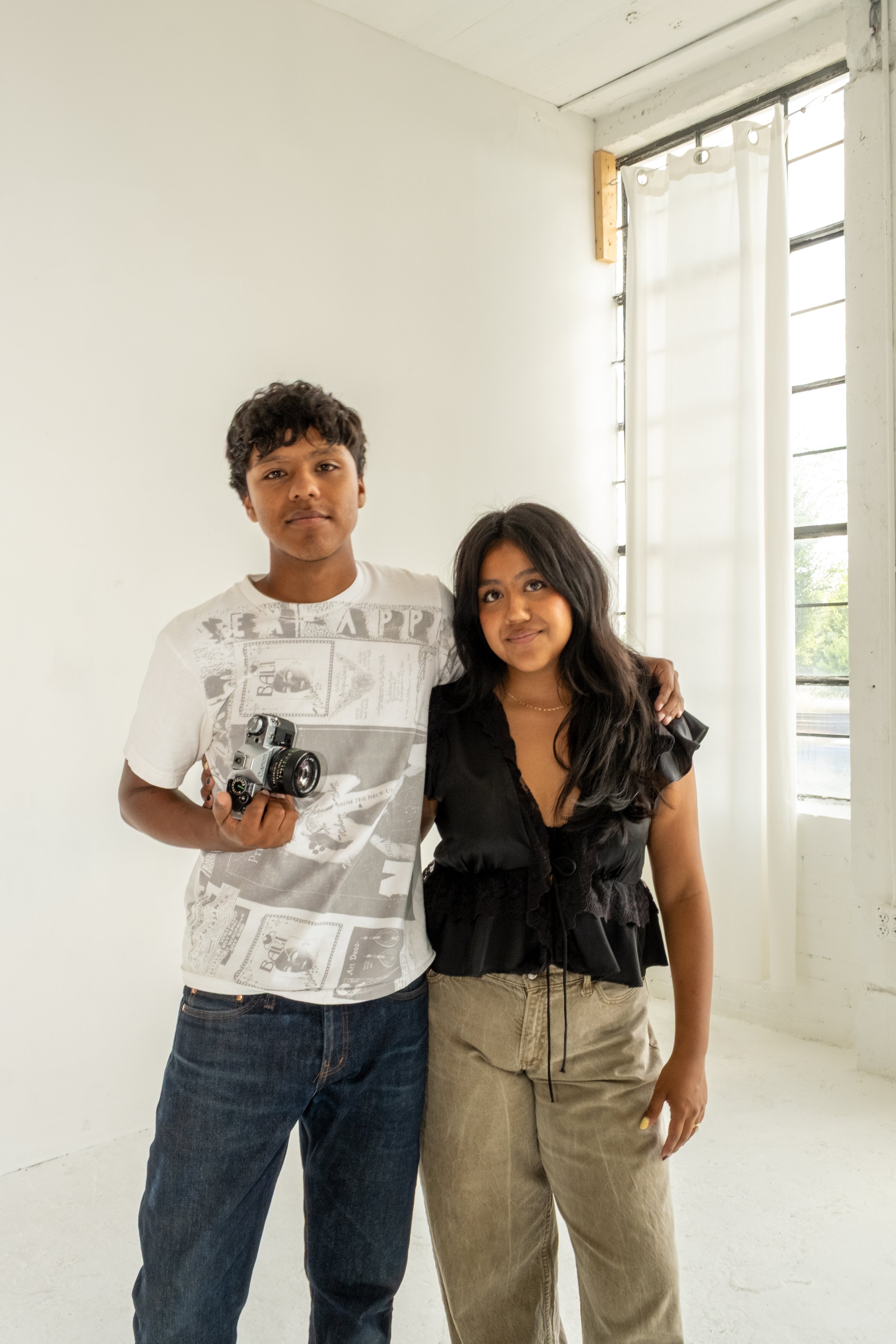 A boy and a girl standing indoors near a window with white curtains. The boy is holding a vintage camera and has his arm around the girl's shoulder. The girl is smiling and wearing a black top and beige pants.