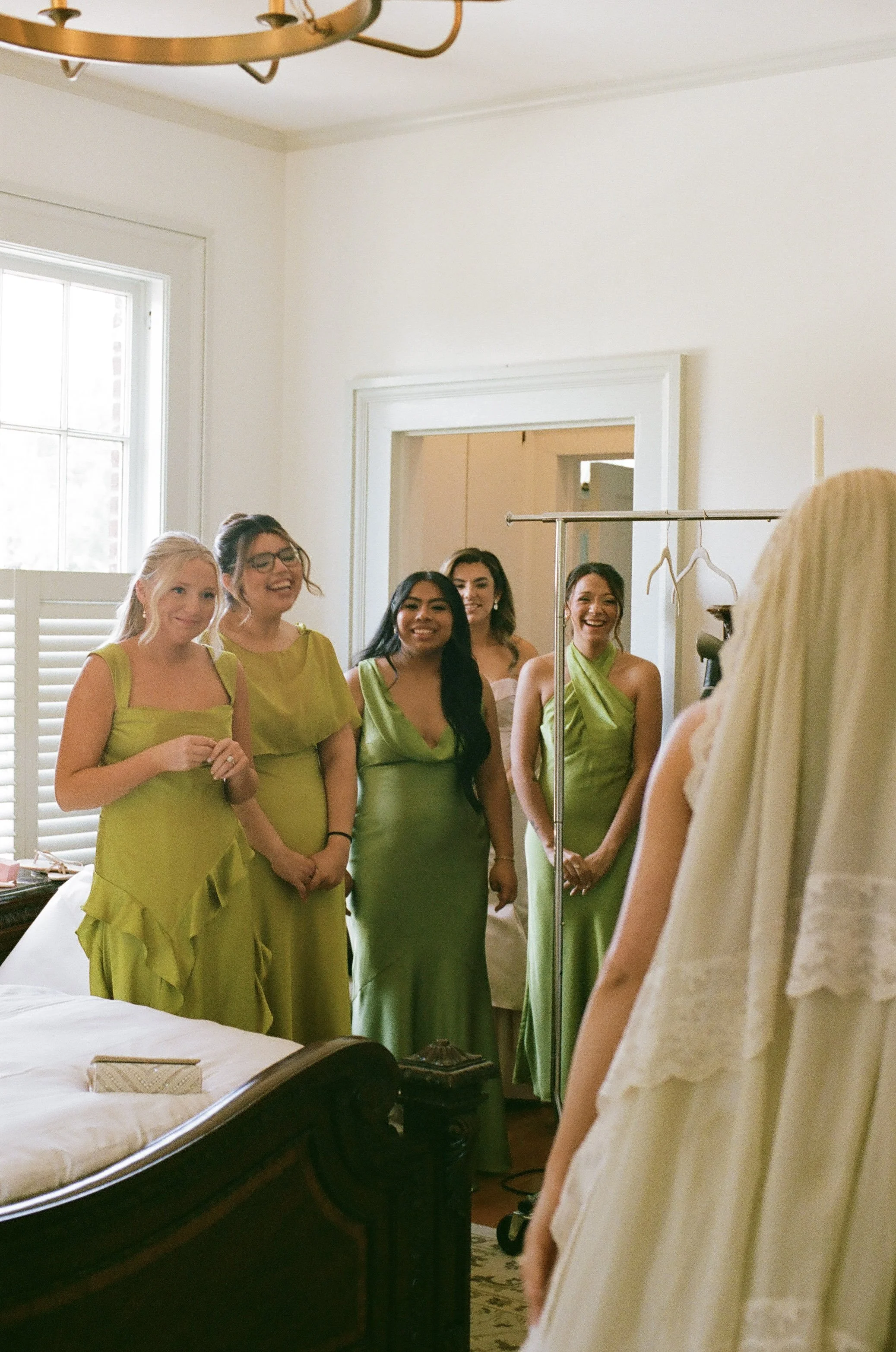 Six women in matching lime green dresses standing in a room, smiling and posing for a photo, with a white bed and window behind them.