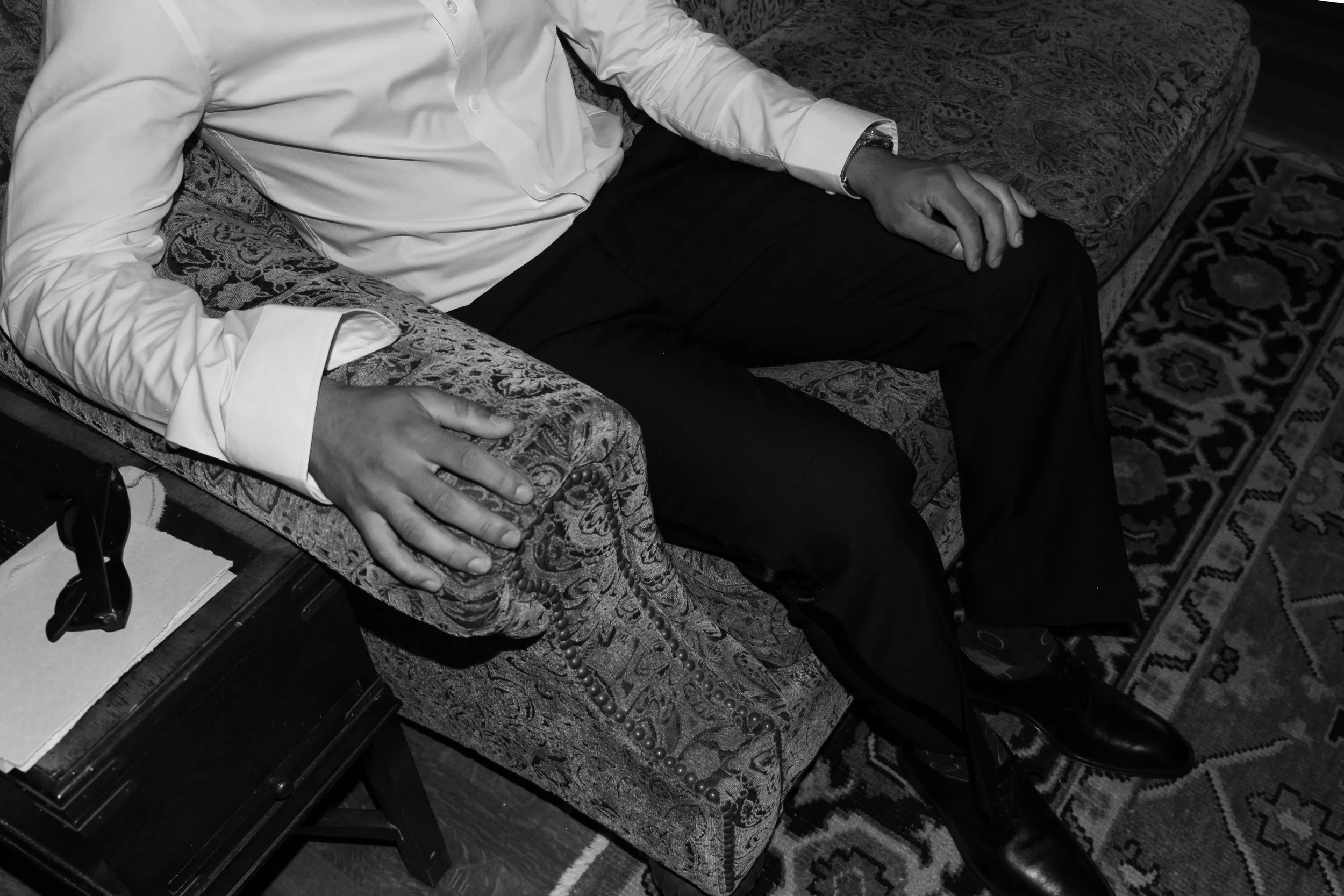 A person wearing a white dress shirt and black pants is sitting on a patterned sofa with their hands resting on their lap. A beaded necklace is draped over the sofa arm. The image is in black and white.