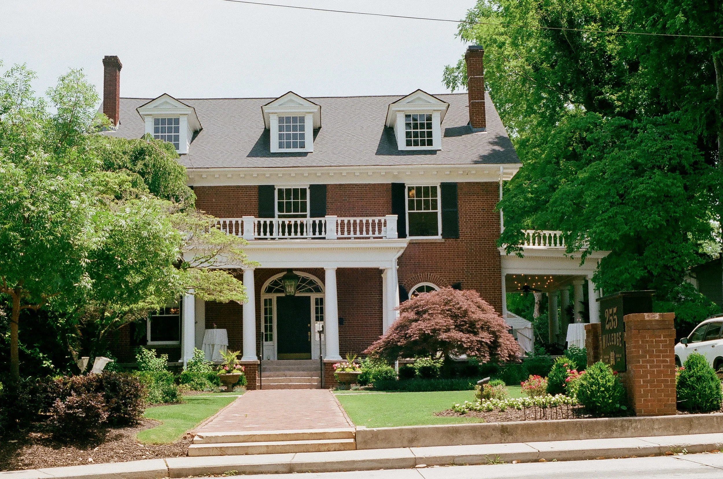 A large brick house with a gray roof and four prominent dormer windows, surrounded by lush green trees and well-manicured bushes, with a brick walkway leading to the front entrance.
