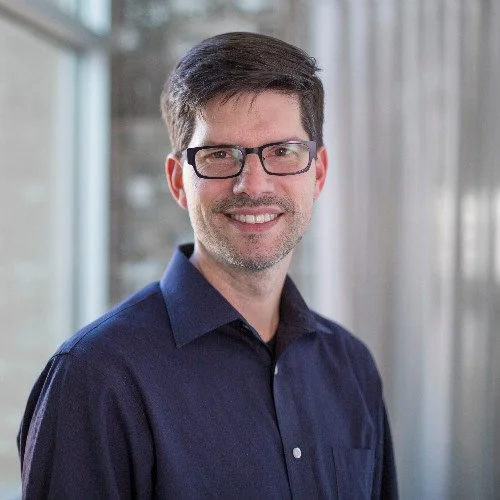 Headshot of a man with glasses, wearing a dark blue collared shirt, smiling in front of a window with curtains.