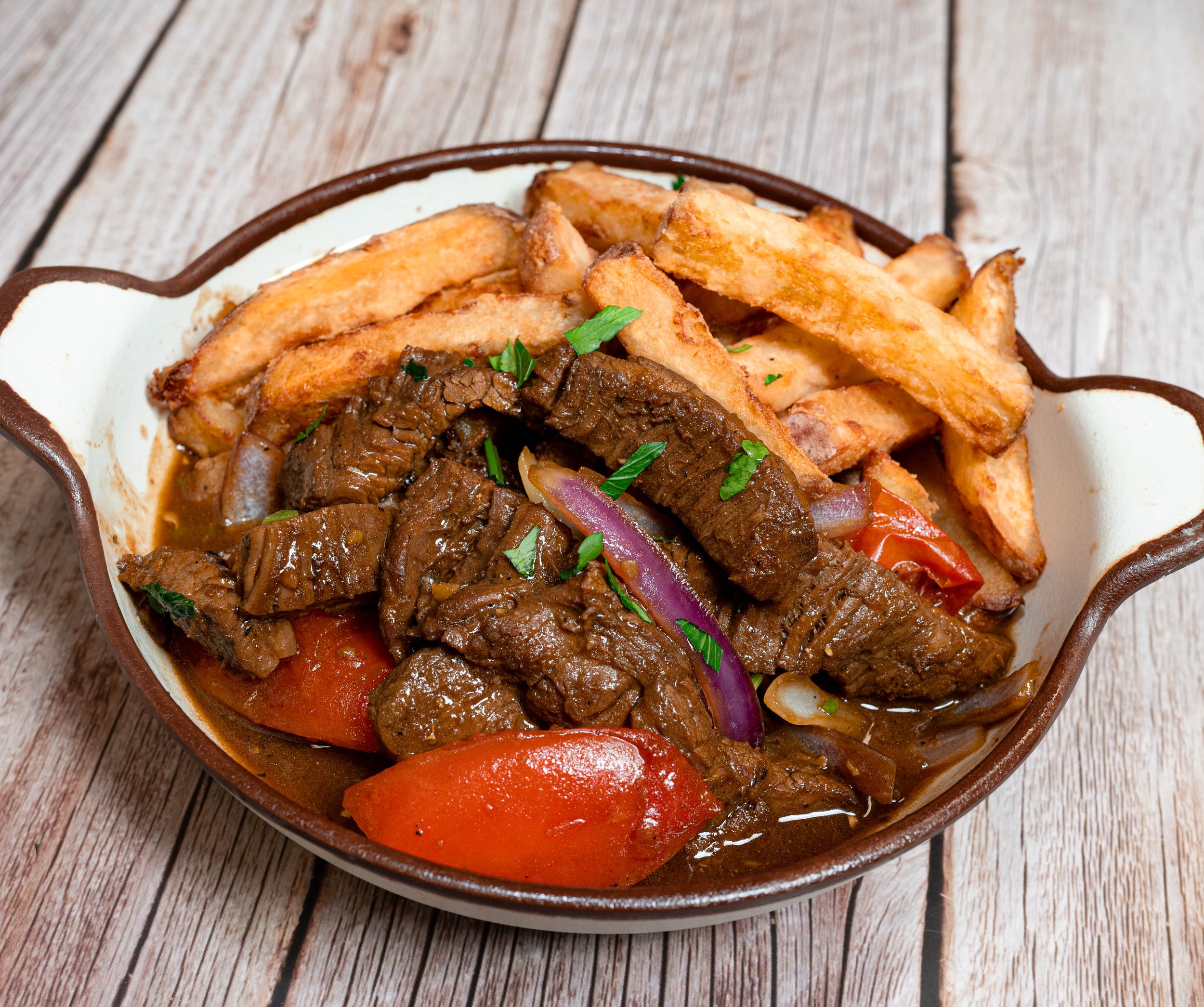 A bowl of Lomo Saltado, a traditional Peruvian dish, featuring sautéed beef strips, red onions, tomatoes, and French fries garnished with parsley on a marbled surface.
