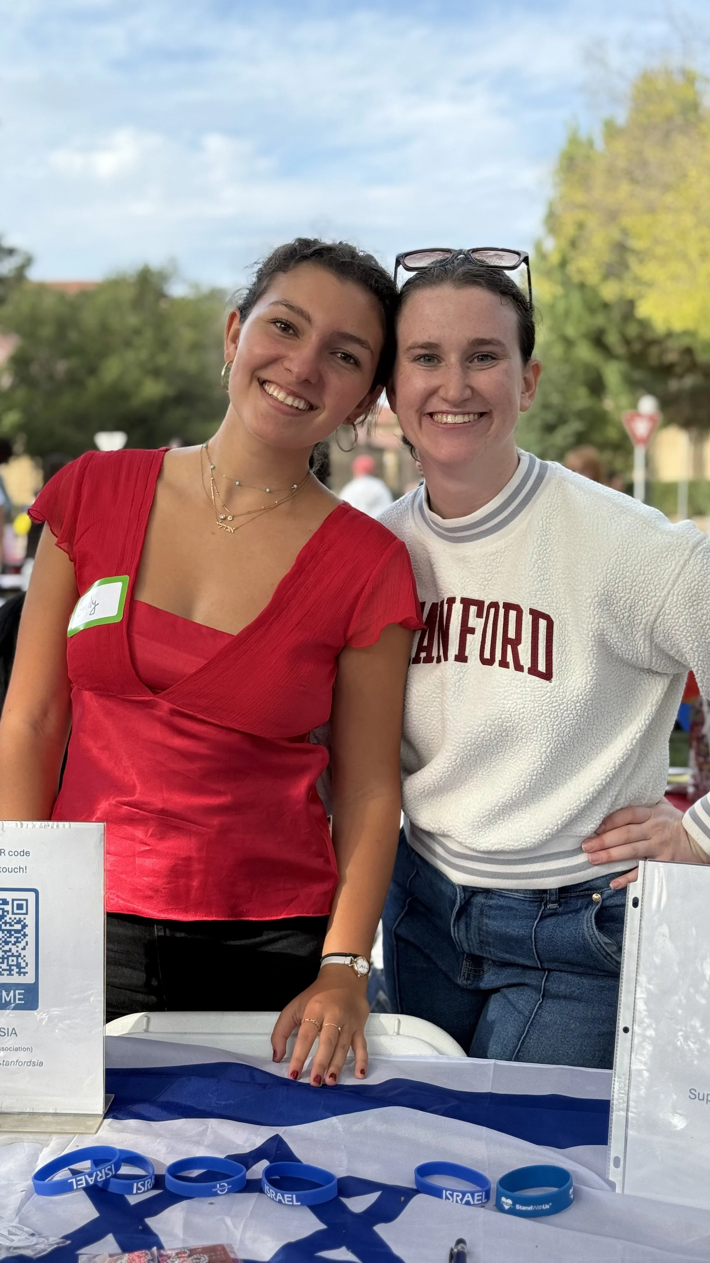 Two smiling women standing behind a table with blue wristbands and a sign, outdoors with trees and a cloudy sky in the background.
