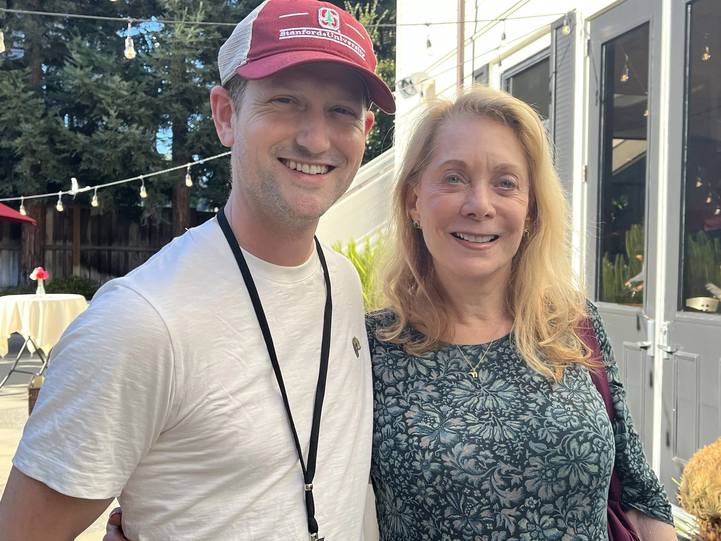 A smiling man wearing a red and white baseball cap with the Stanford University logo and a white t-shirt, standing next to a smiling woman with long blonde hair wearing a floral top outdoors in a backyard setting with string lights and a food truck in the background.