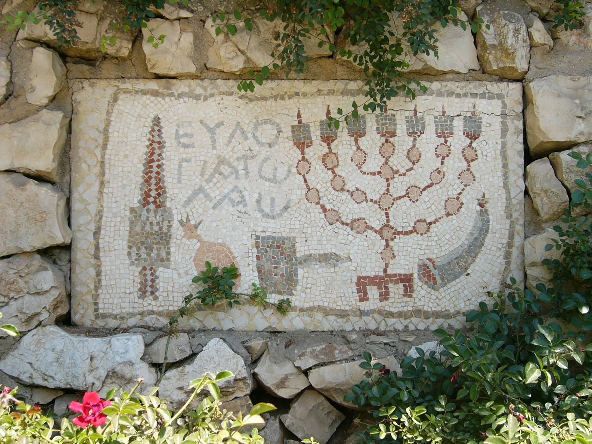 A mosaic with a menorah, a cat, and Hebrew text, surrounded by rocks and green foliage.