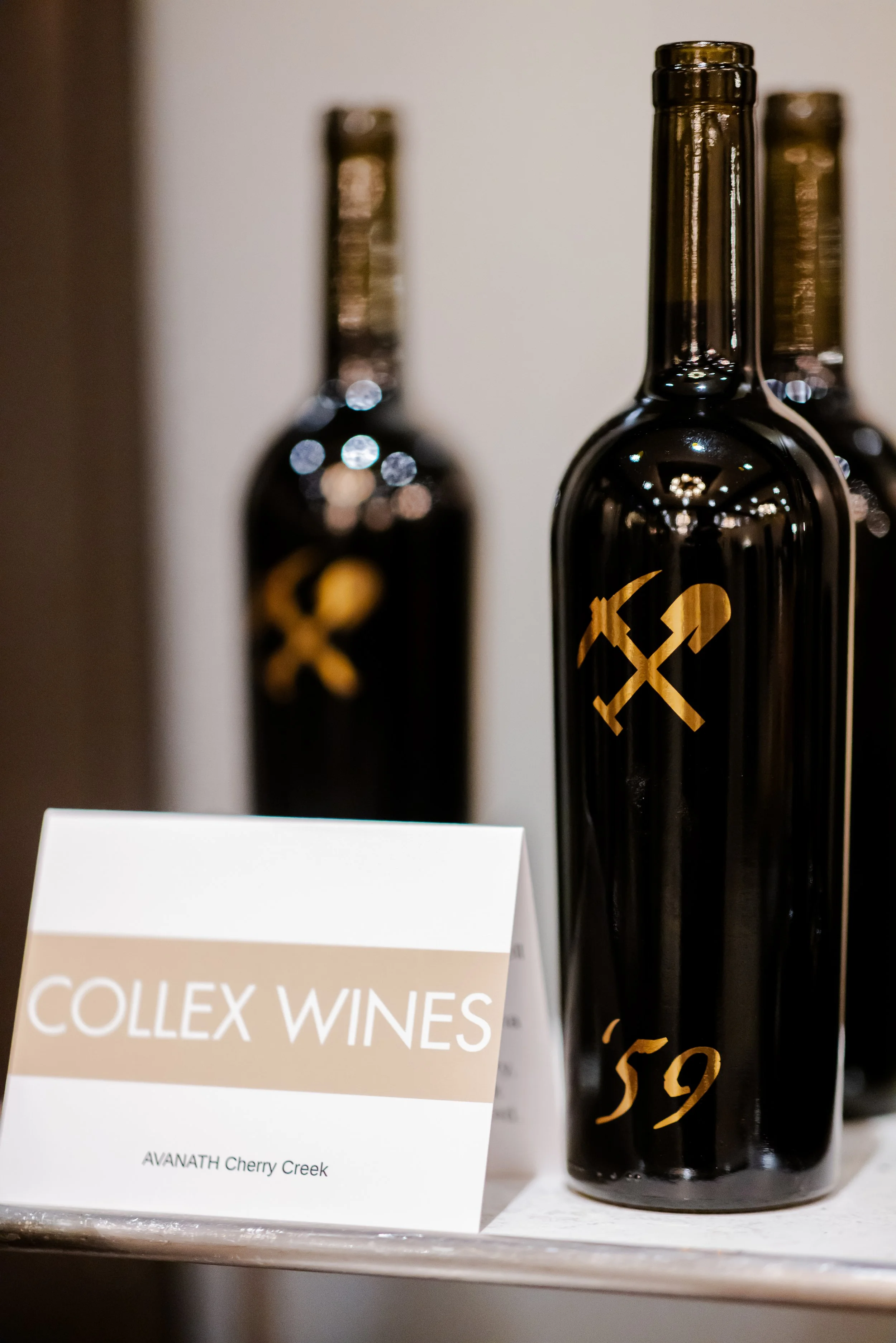 Black wine bottles with gold symbols on a display shelf, a sign reading 'COLLEX WINES AVANATH Cherry Creek' in front of the bottles.