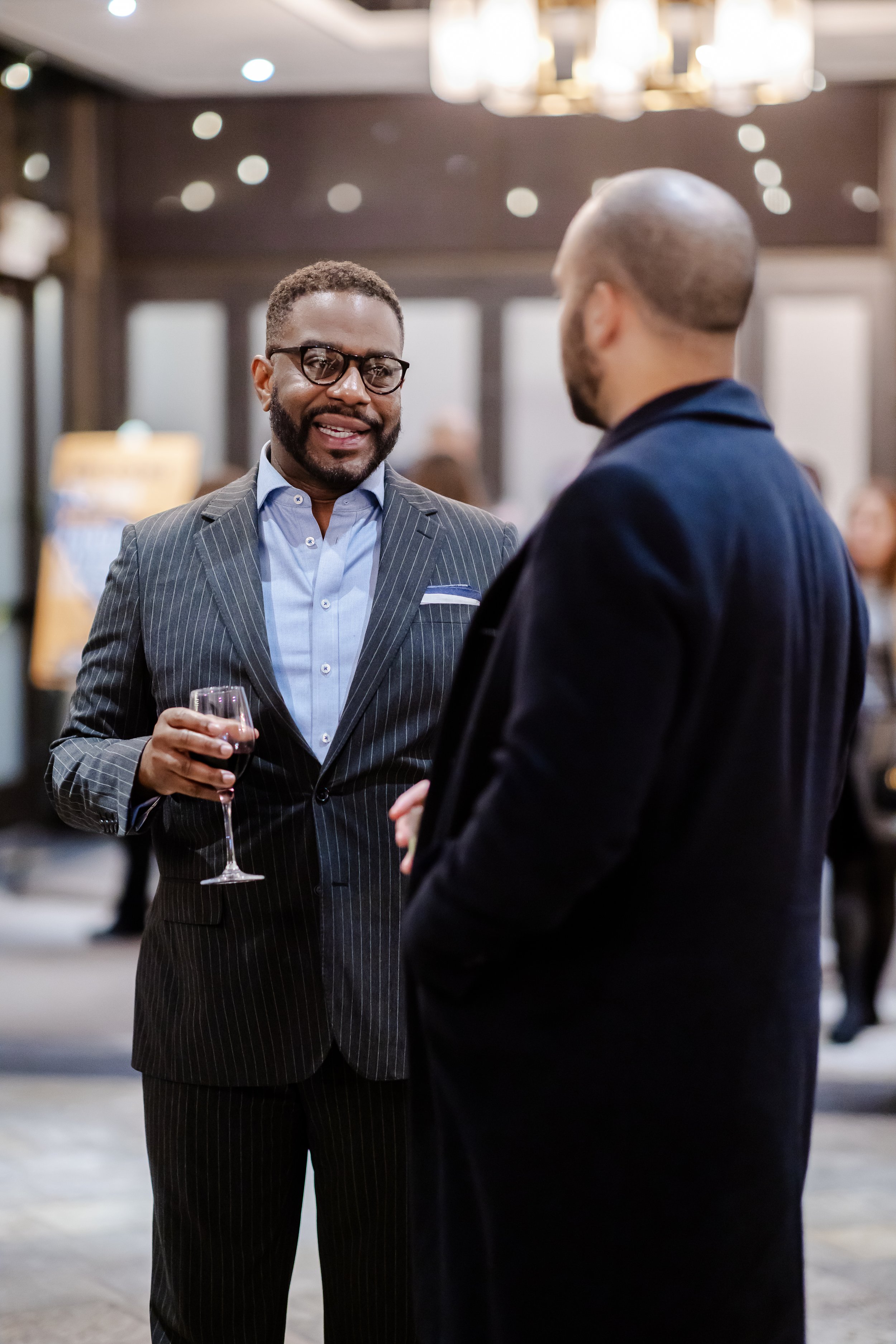 Two men in suits having a conversation at a social event. One is holding a glass of red wine.