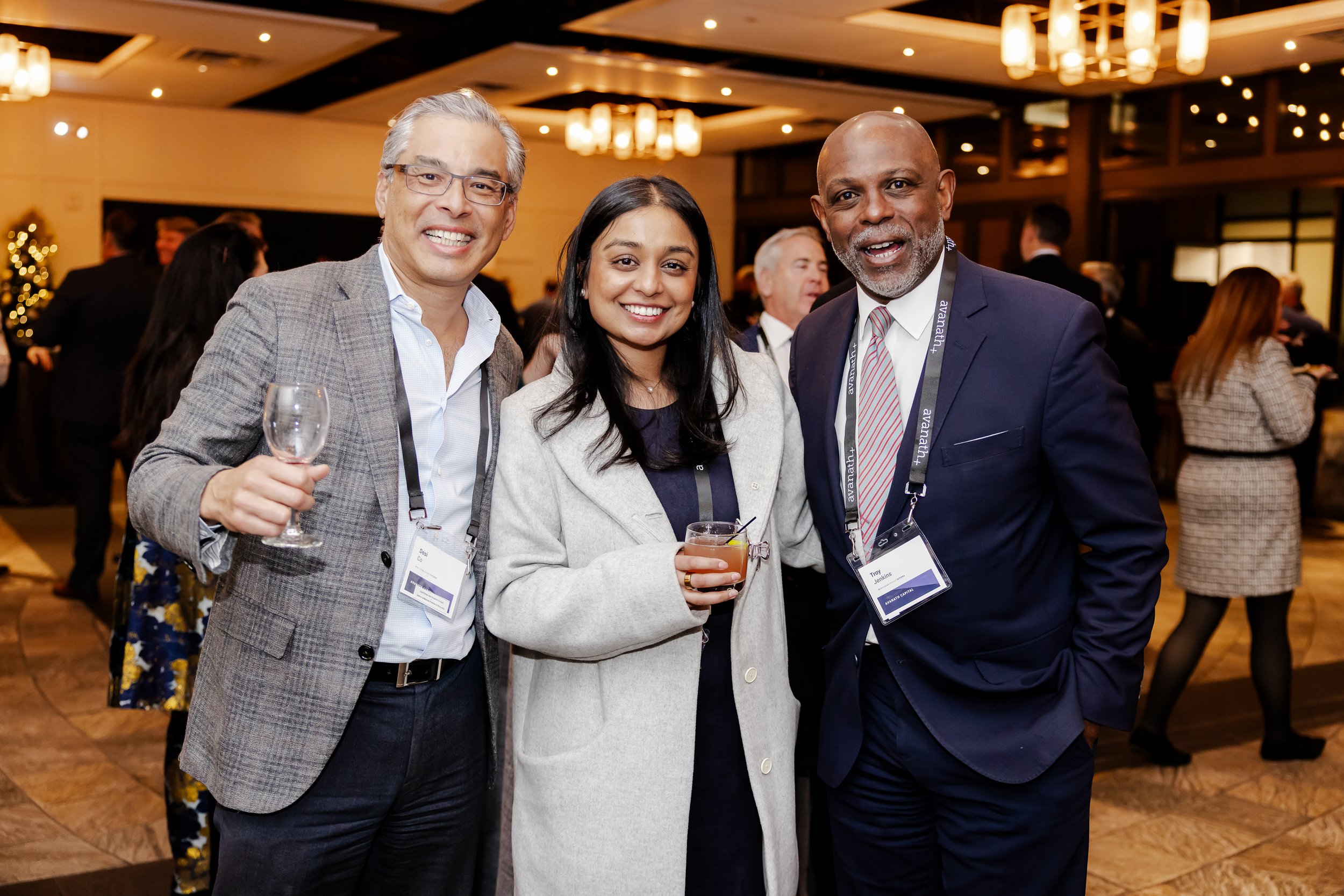 Three people at a professional networking event, smiling for the camera with drinks in hand, in a well-lit, modern venue with other attendees in the background.