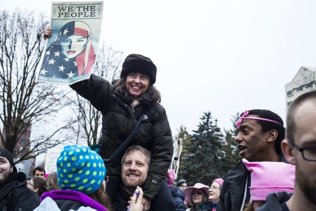   Monica Silva, 49, of East Lansing, Mich., sits on her husband’s shoulders during the Women’s March on Lansing outside of the Michigan State Capitol Building in Lansing, Mich., on Saturday, January 21, 2017. (Photo by Brittany Greeson/GroundTruth)  