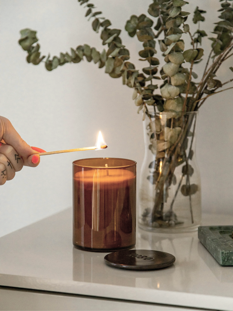 A woman's hand lighting a candle with eucalyptus in the background in a vase helping create a peaceful environment for a new cancer patient, a caregiver, parent or survivor