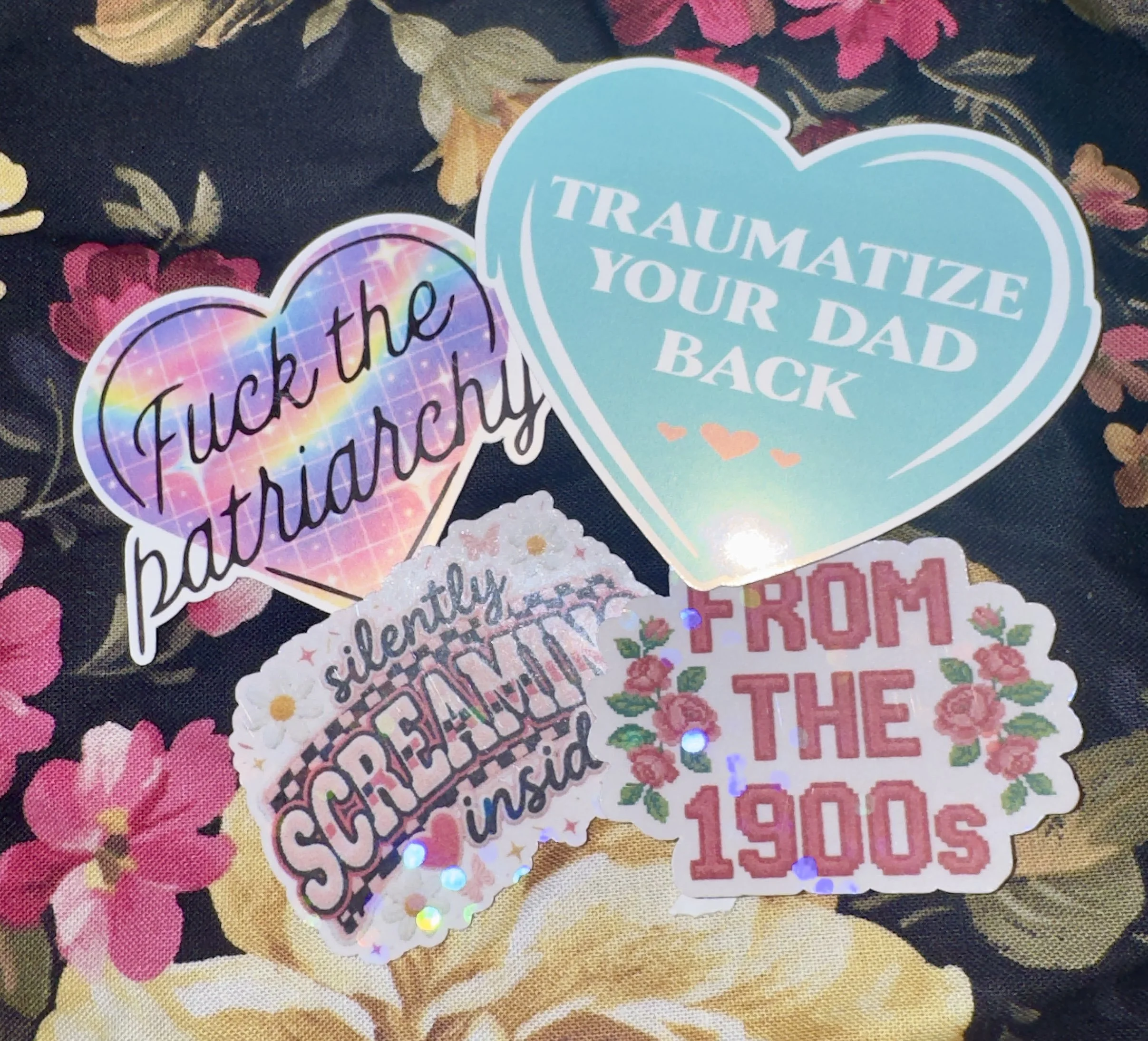 Close-up of several colorful stickers with positive messages on a floral-patterned fabric background. The stickers have phrases like 'Fuck the Hatred,' 'Traumatize Your Dad Back,' and other uplifting or expressive words, some with decorative fonts and holographic effects.