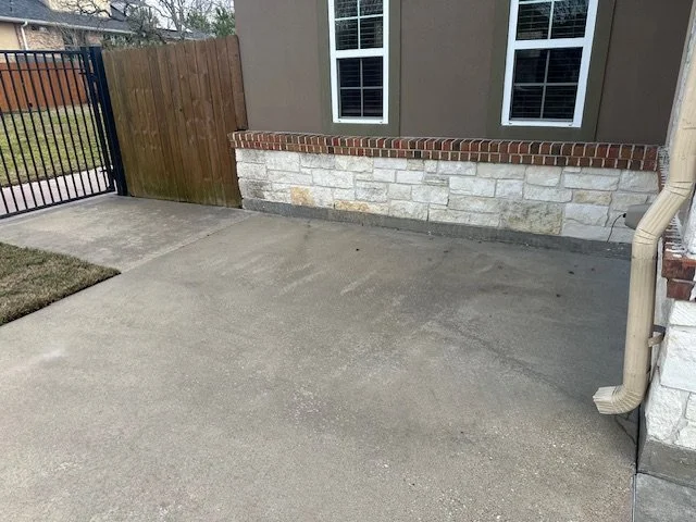 Concrete patio area next to a house with two windows, a brick wall, and a wooden fence with a gate.