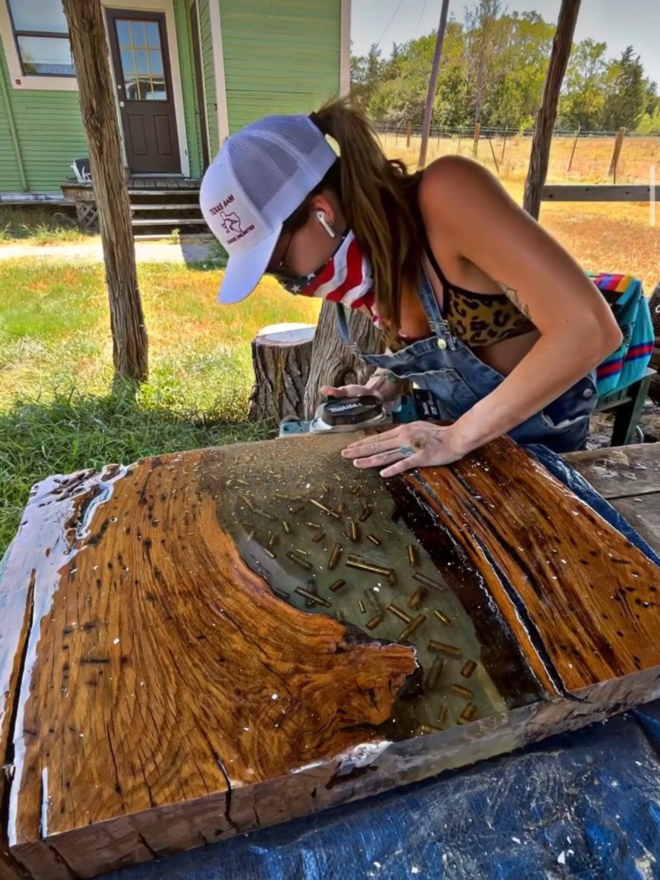 A woman wearing a trucker hat, leopard print top, overalls, and a mask with the American flag design is working on a large wooden piece, possibly a wood carving or epoxy table, outdoors under a canopy. She is using a tool while focusing on the wooden art piece.