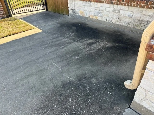 A freshly paved black asphalt driveway next to a house with stone and brick exterior, a metal gate at the entrance, and a small patch of grass.