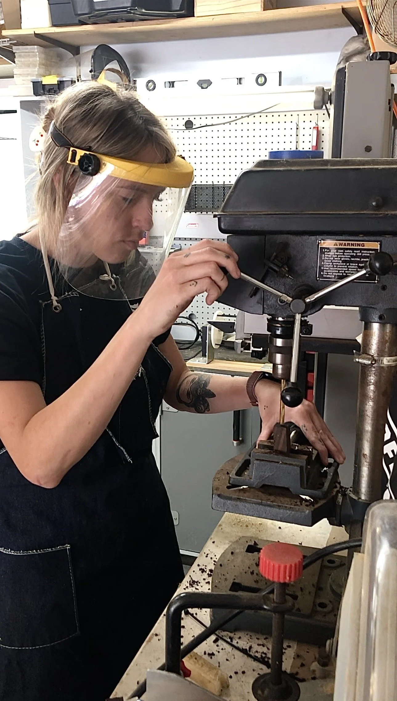 A woman wearing a face shield and black apron working at a drilling machine in a workshop.
