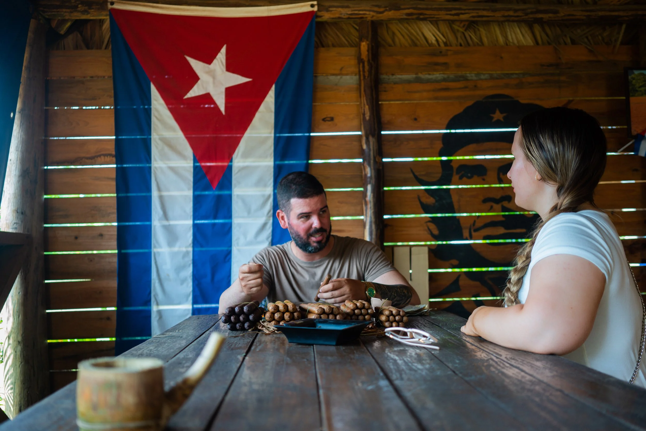 Two people sitting at a wooden table inside a rustic room with a Cuban flag and a portrait of Che Guevara on the wall, having a conversation.