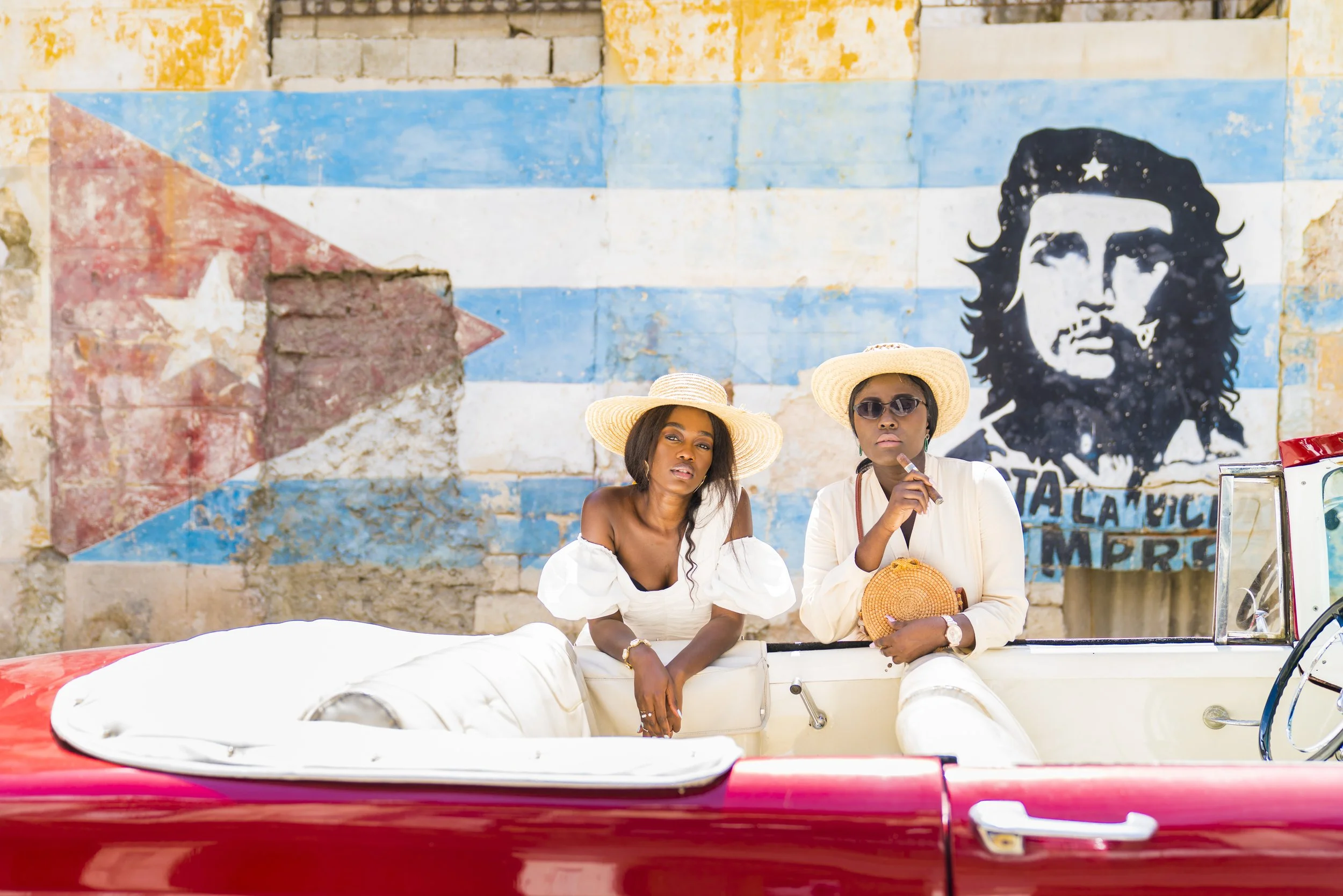 Two women sit in a vintage red convertible car in front of a weathered mural of Che Guevara with blue and white stripes and a red triangle with a white star, with a partially crumbling brick wall background. Both women wear white blouses, sunglasses,