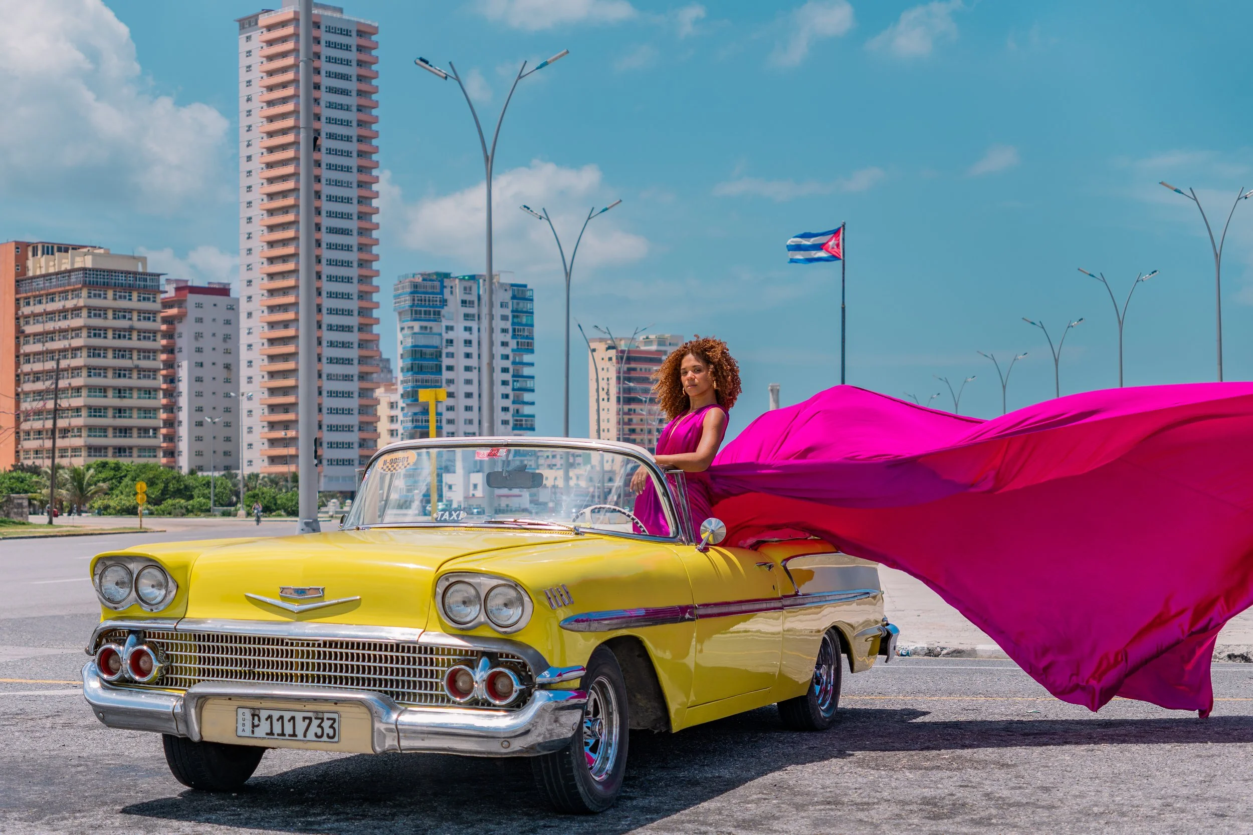 A woman in a pink dress standing in a vintage yellow Cadillac convertible with a large pink fabric flowing from the back, parked in an urban area with tall buildings, streetlights, and a Cuban flag in the background.