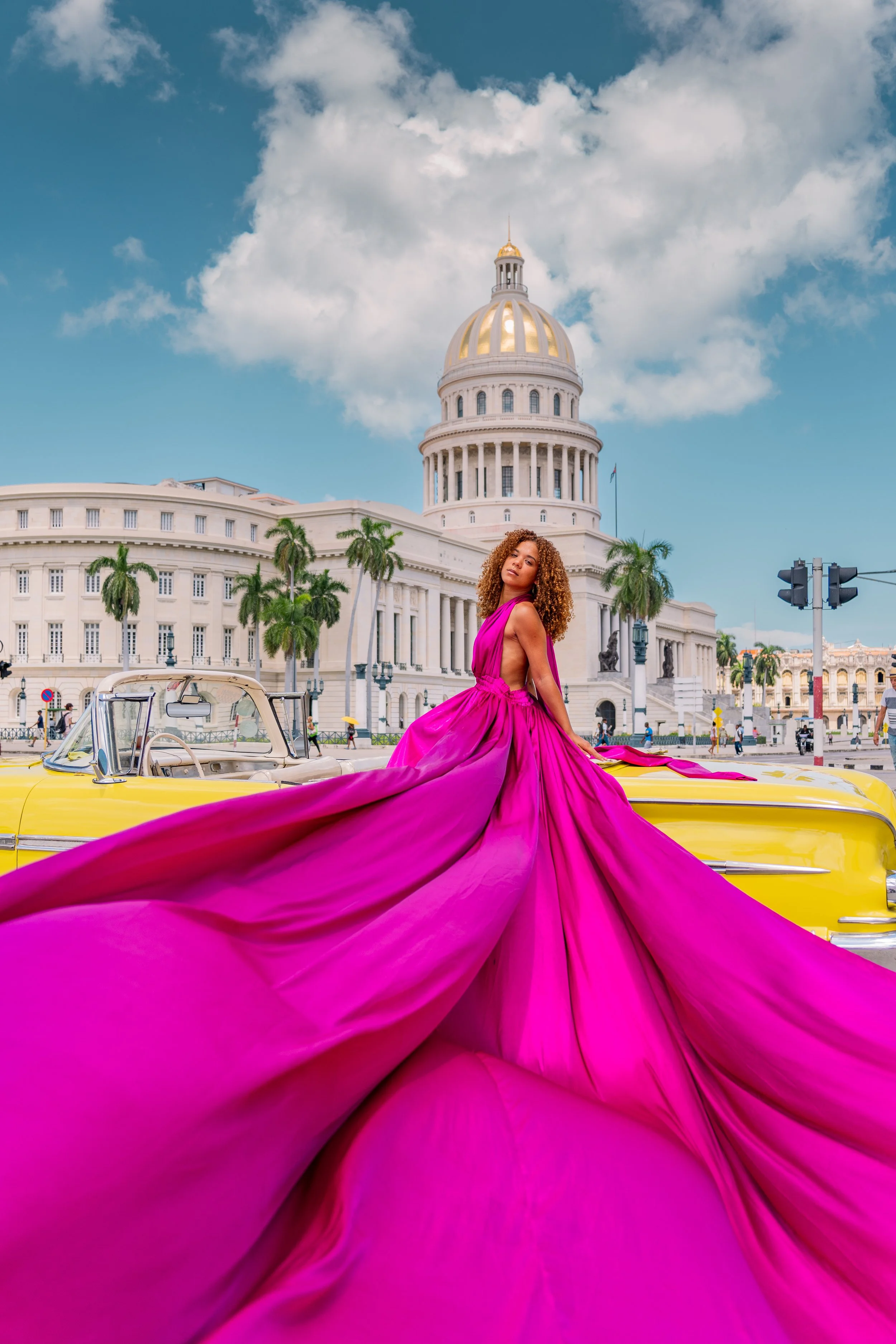 A woman in a vibrant fuchsia gown standing in front of a yellow vintage car in front of the Capitol Building in Havana, with palm trees and a partly cloudy sky in the background.