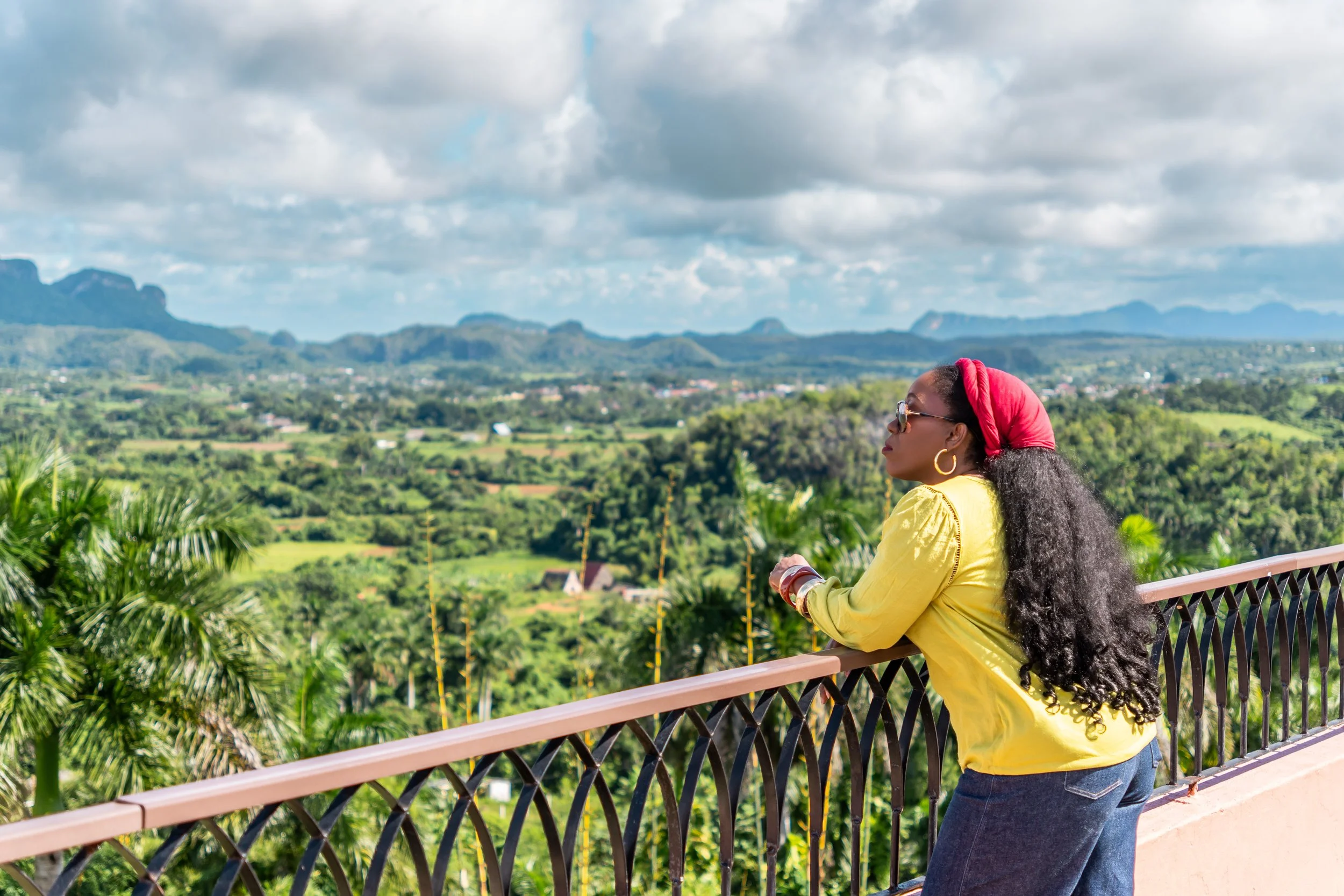 A woman with long curly hair, wearing a red headscarf, sunglasses, a yellow top, and blue jeans, leaning on a railing and overlooking a green valley with mountains in the background in Viñales, Cuba.