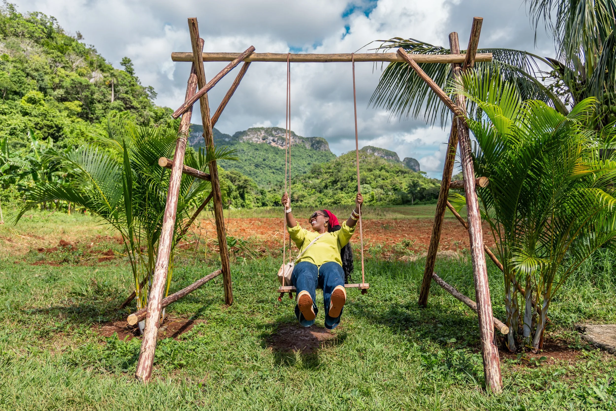 A woman sitting on a wooden swing in a lush green outdoor setting with mountains in the background in Viñales, Cuba.