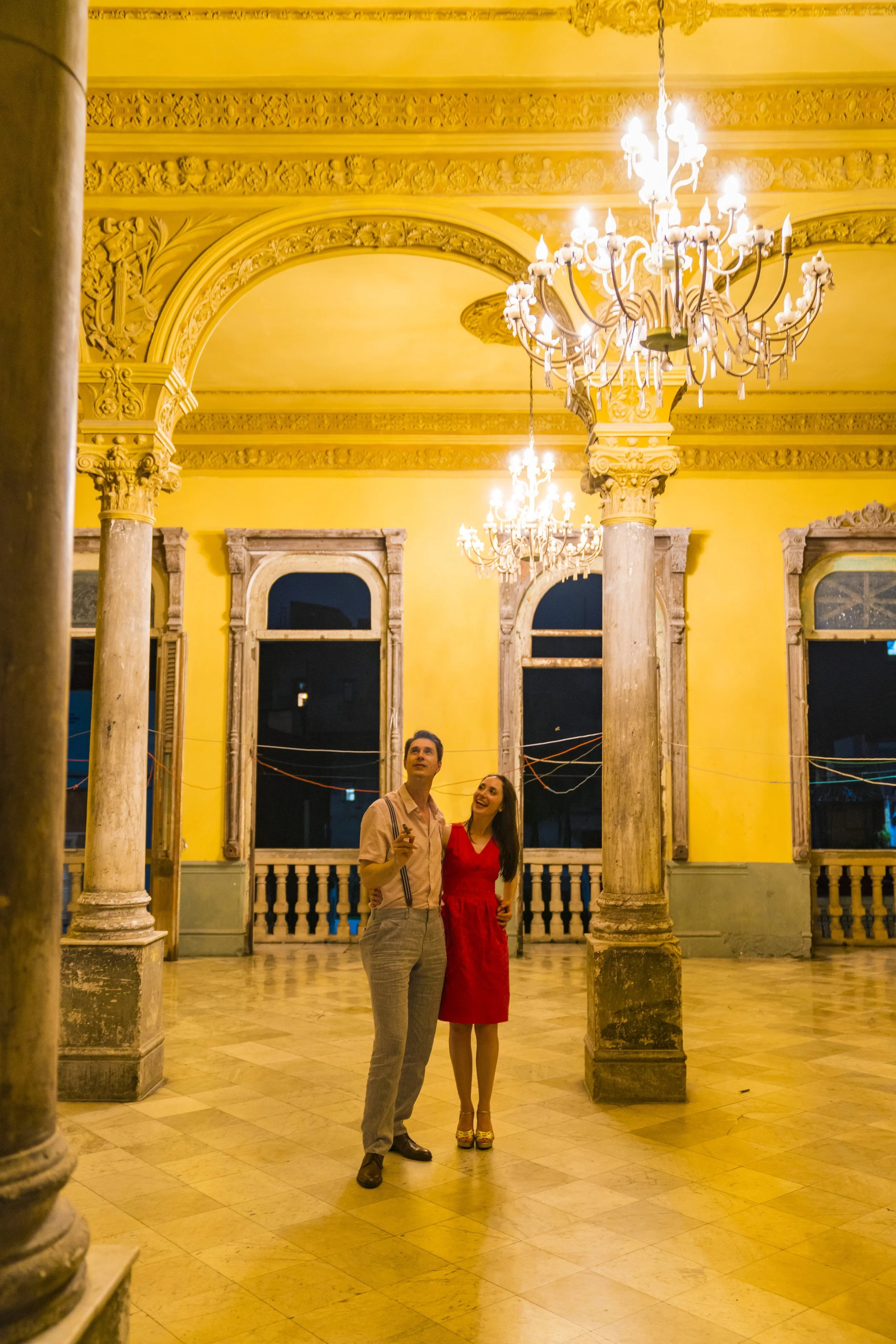 A man and woman stand inside an ornate, yellow-walled room with chandeliers and tall windows, looking up and smiling.