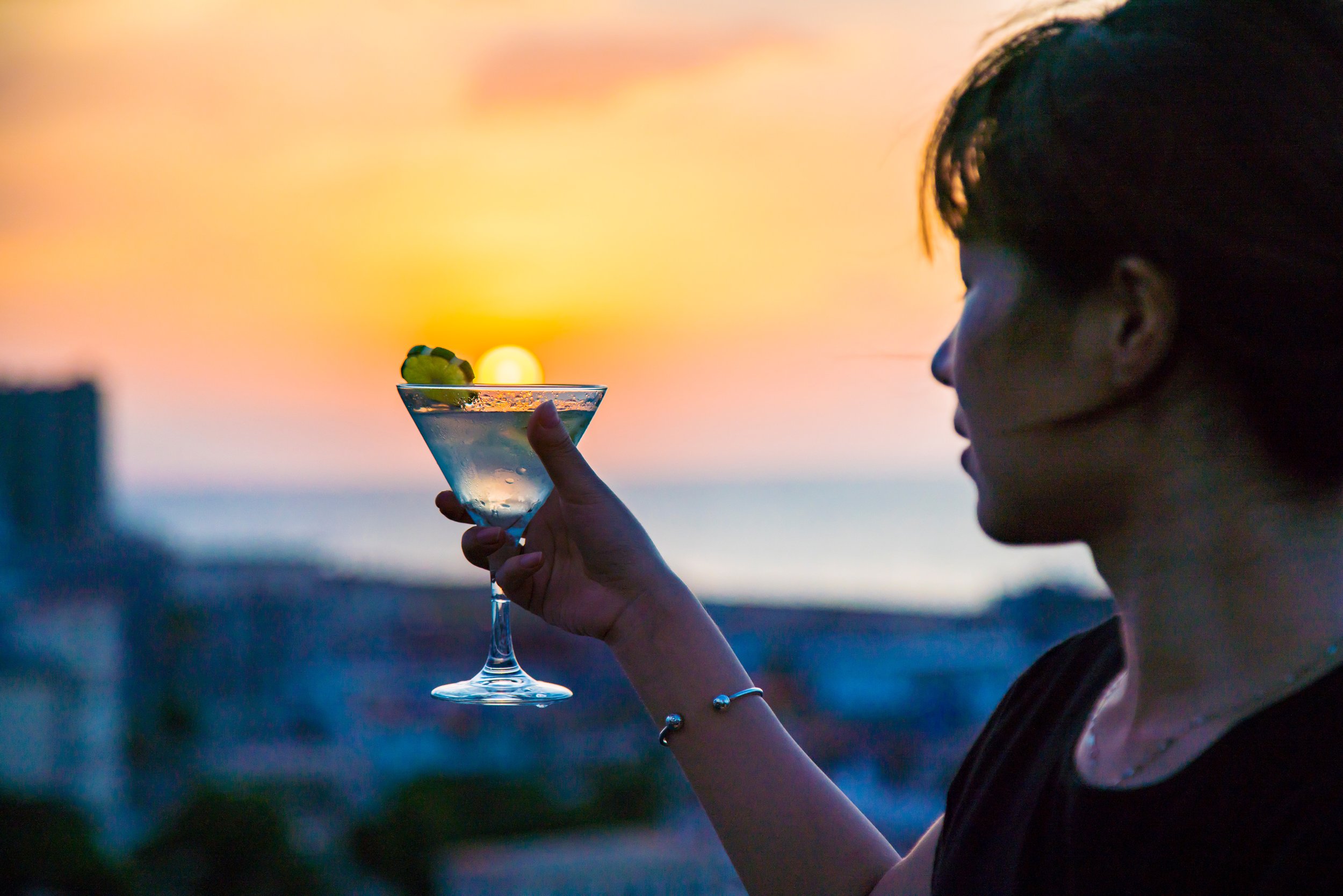 A woman holding a martini glass with a lime wedge, overlooking a sunset over the ocean.