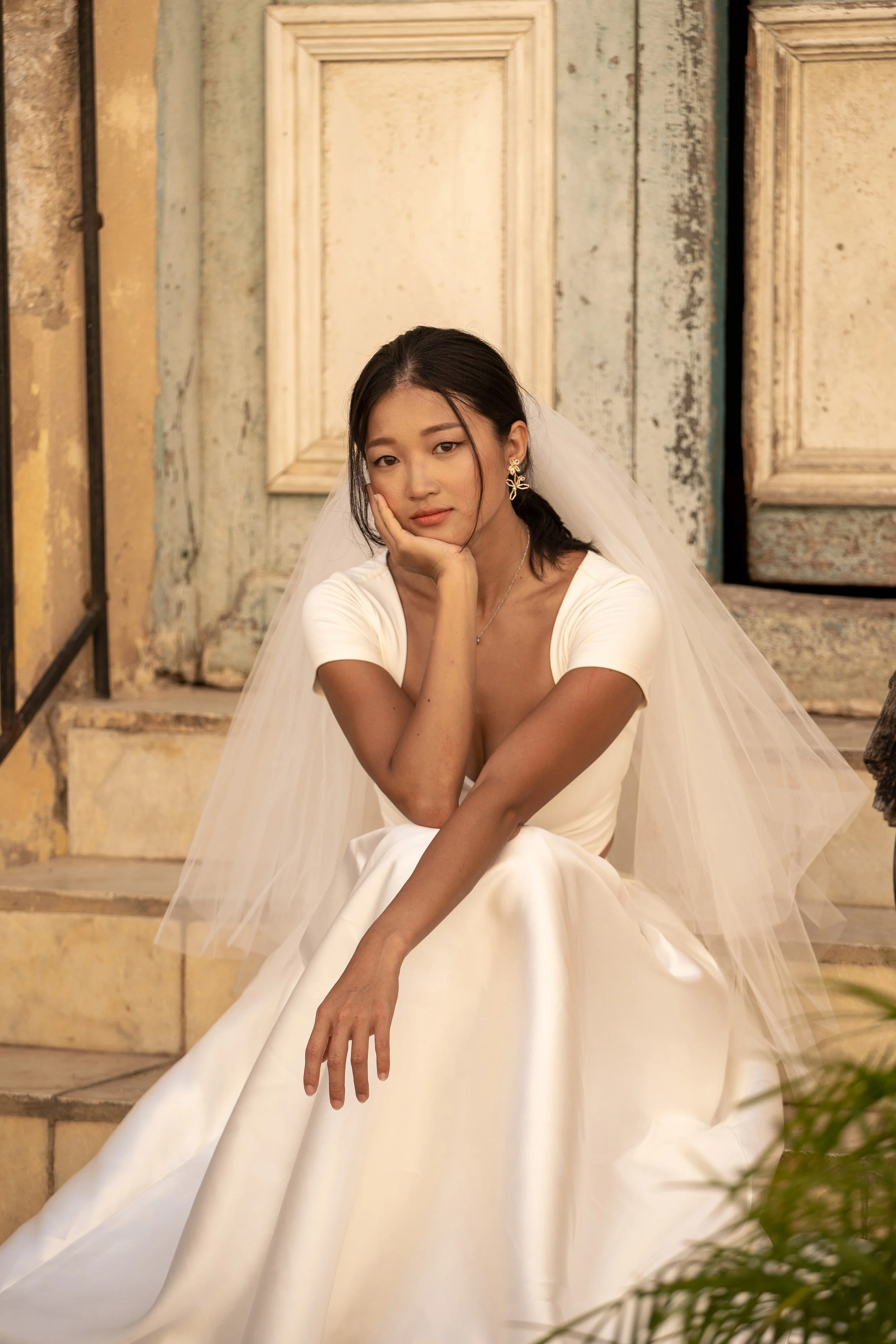 Bride sitting on steps in front of an old door, wearing a white wedding dress and veil.