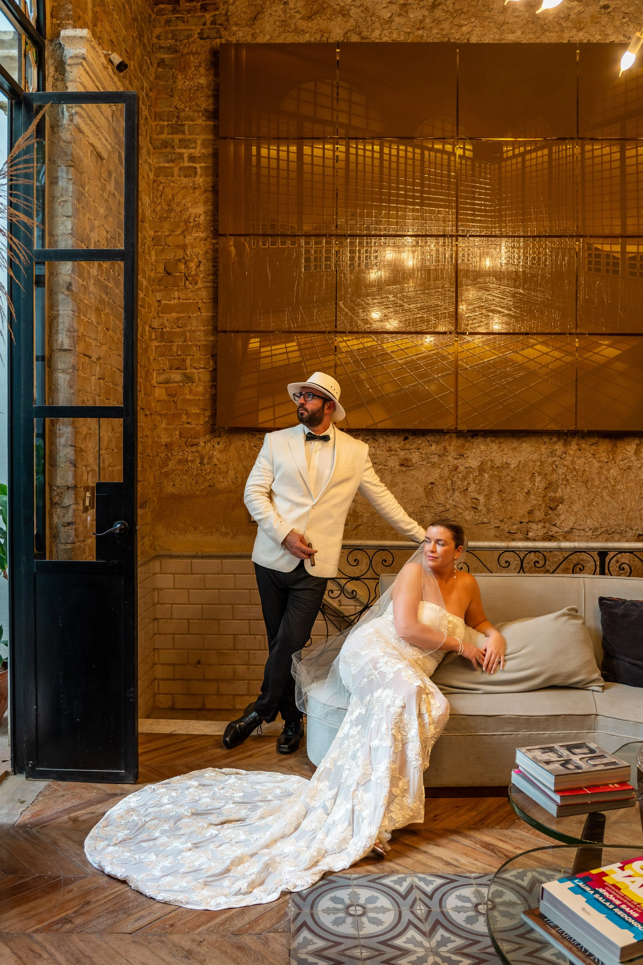 A bride in a white wedding dress and veil is sitting on a sofa, while a groom in a white tuxedo jacket, black pants, and a white hat stands beside her with one hand on the sofa. The setting is an indoor space with exposed brick walls and modern art o