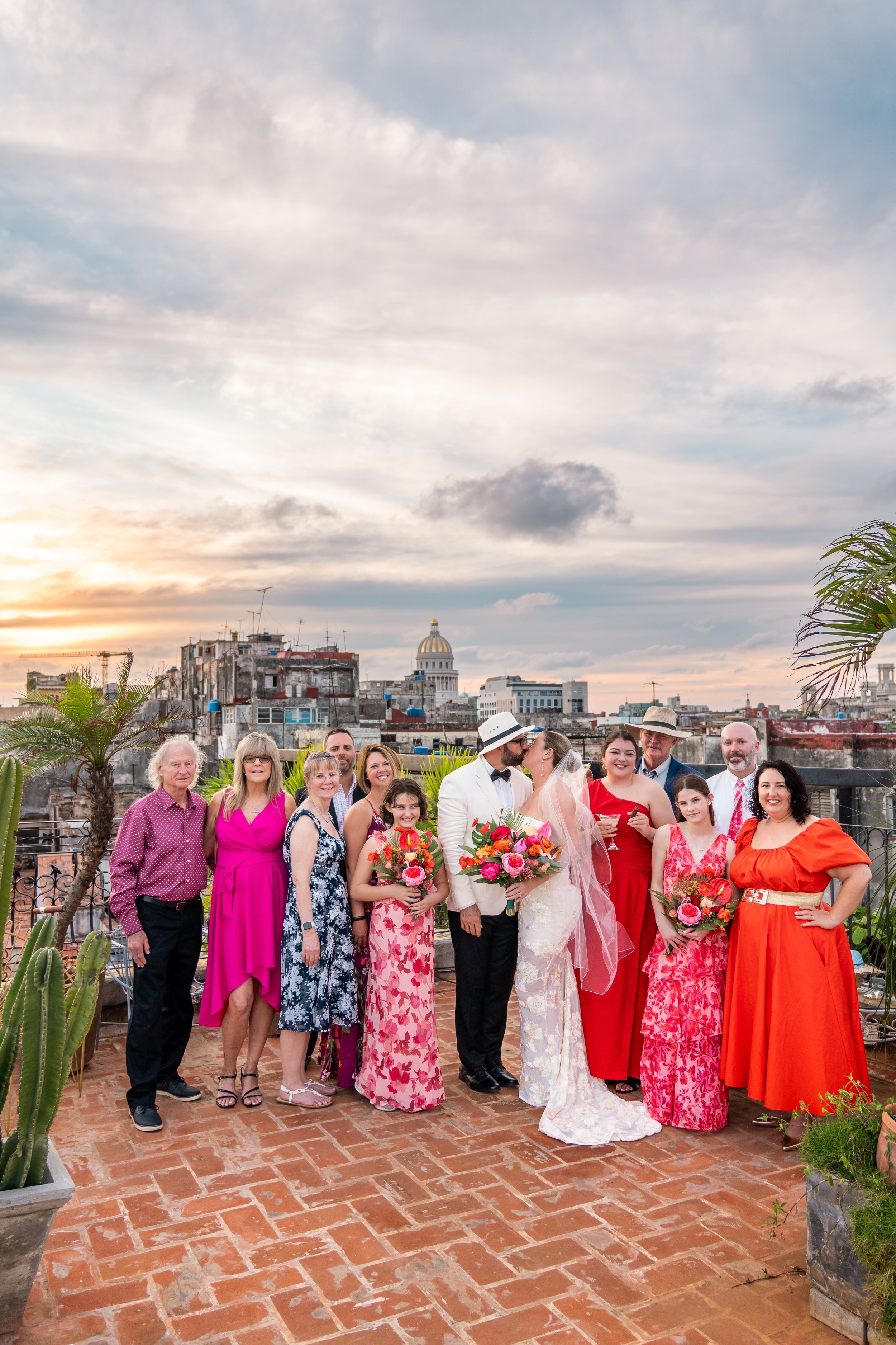 A group of people at a wedding celebration on a rooftop terrace during sunset, with city buildings and a cloudy sky in the background. The bride and groom are kissing, holding bouquets, surrounded by family and friends in colorful attire.