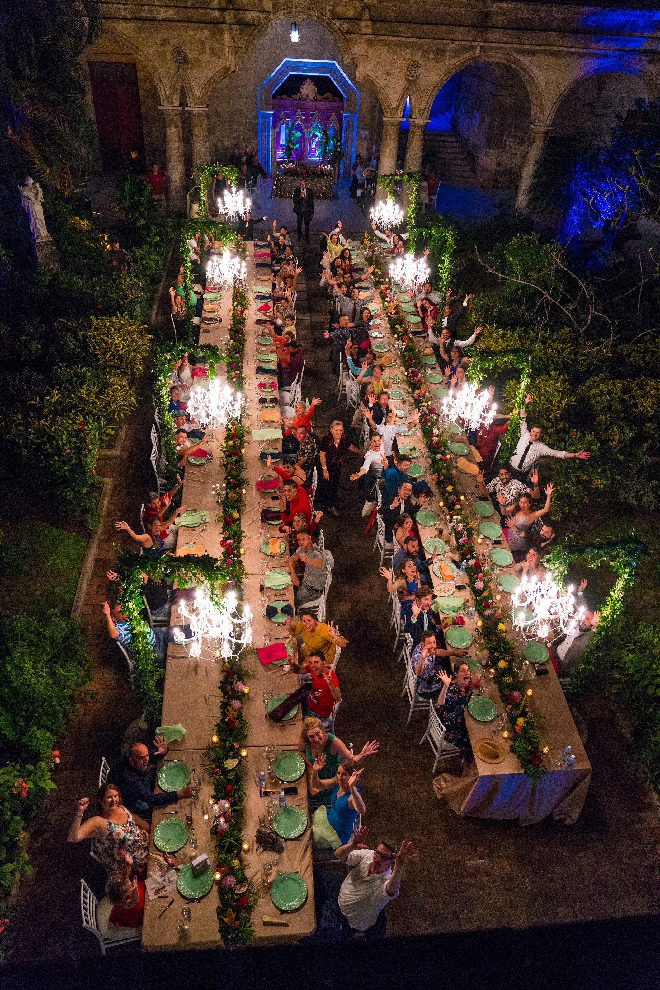 Aerial view of a festive outdoor dining scene with long tables set for a celebration. People are sitting and standing around the tables, waving and smiling. The tables are decorated with flowers and illuminated by chandeliers. The setting appears to be in a historic courtyard with stone arches.