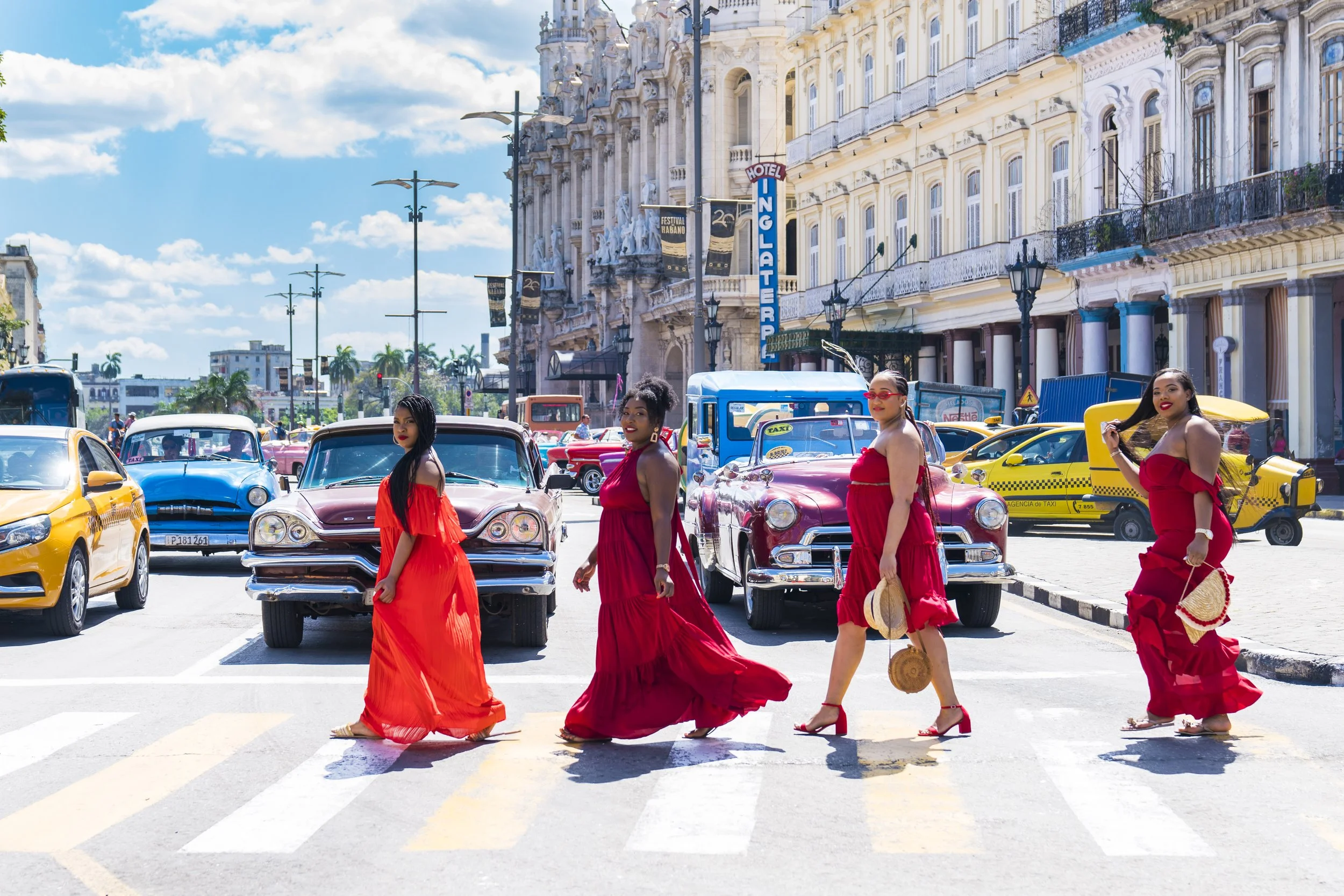 Four women dressed in red dresses crossing a street in a city with vintage cars and colorful taxis parked along the road. The background features ornate buildings, street lamps, and a sign for hotel Inglaterra on a sunny day.