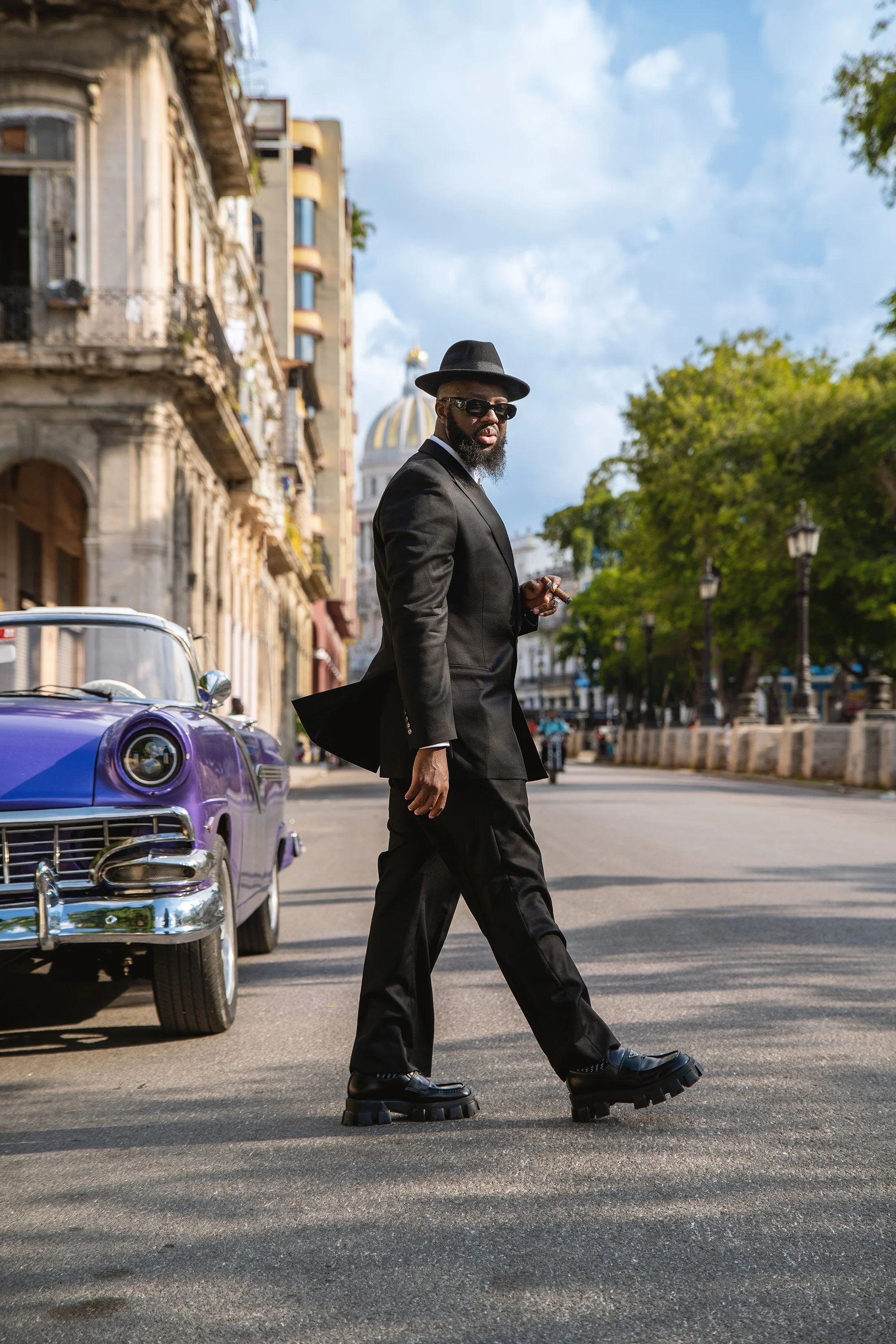 Man in a black suit and hat walking on a city street, with a vintage purple car and historic building in the background.