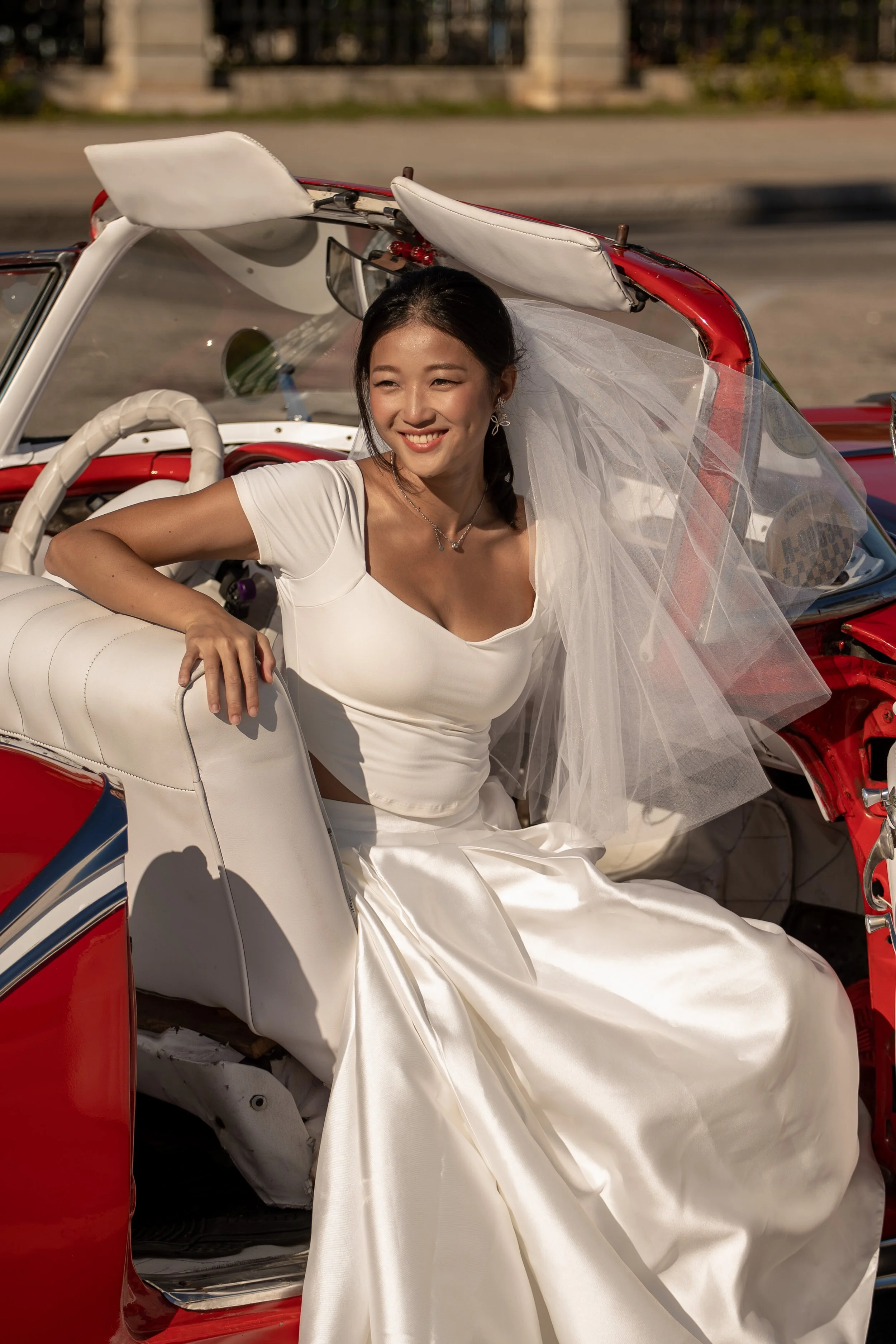 A woman in a white wedding dress and veil sitting inside a vintage red convertible car.
