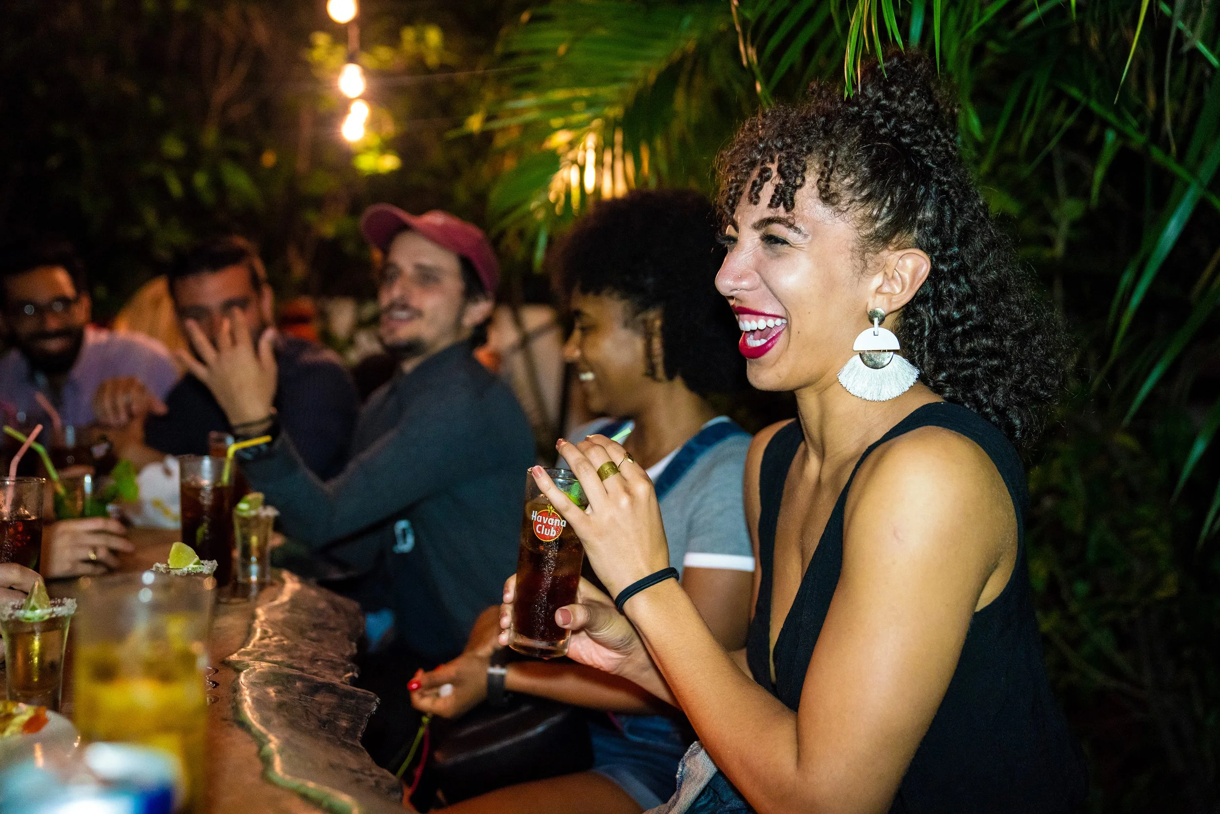 Group of friends enjoying drinks and conversation at a nighttime outdoor gathering, surrounded by lush greenery.