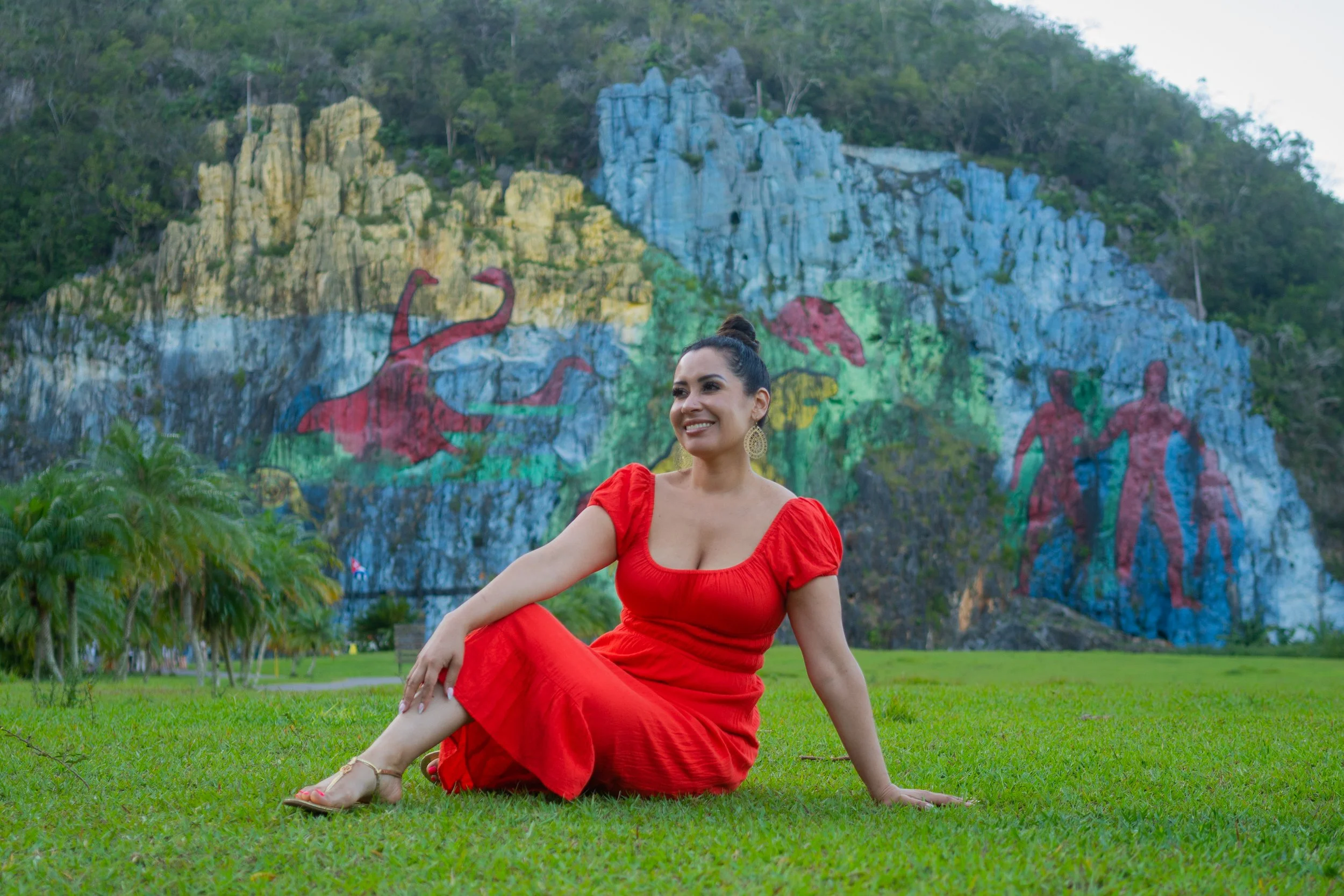 A woman in a red dress sitting on green grass in front of a rocky hill with colorful painted figures and designs.