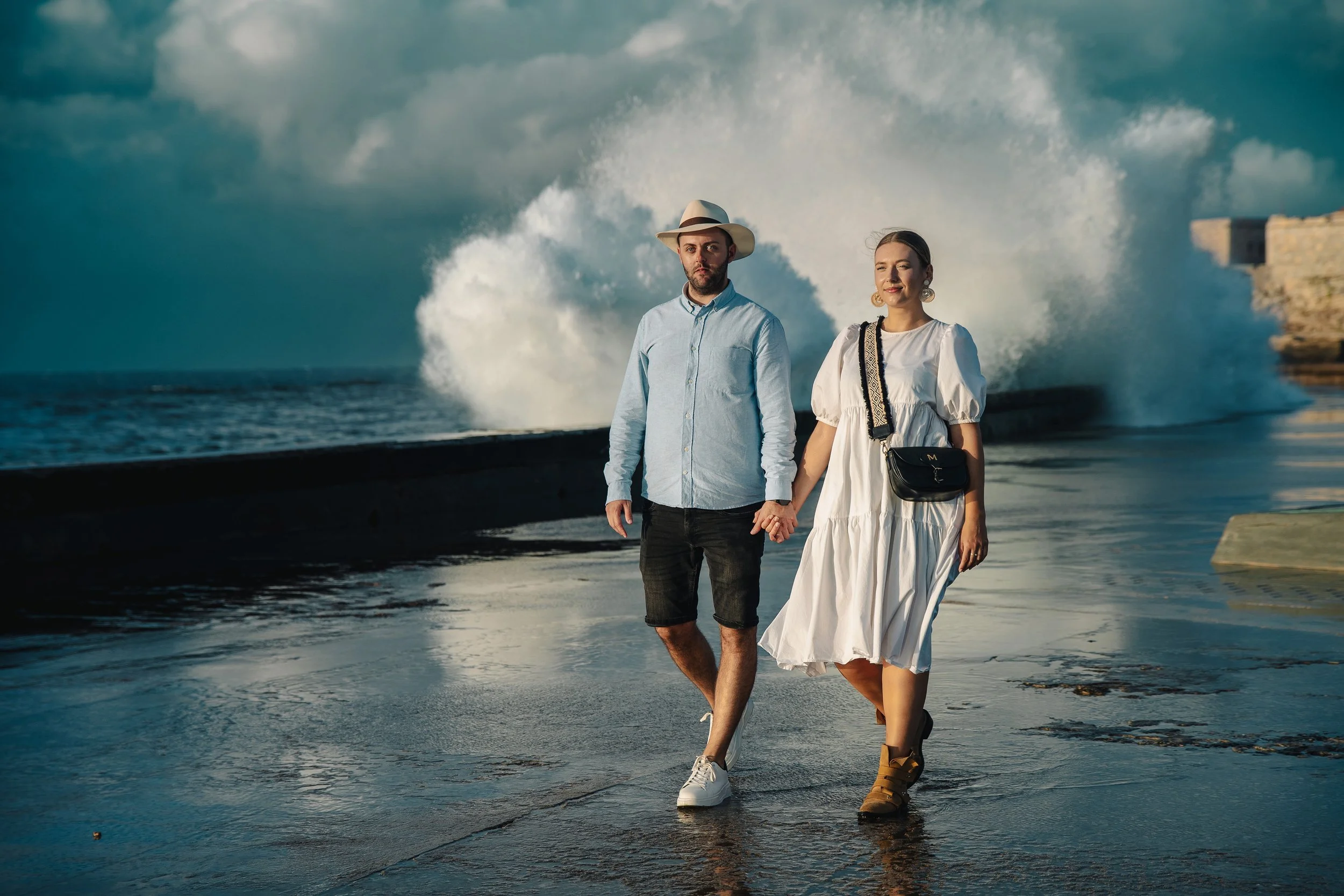 Couple walking along ocean promenade with large waves crashing in the background.