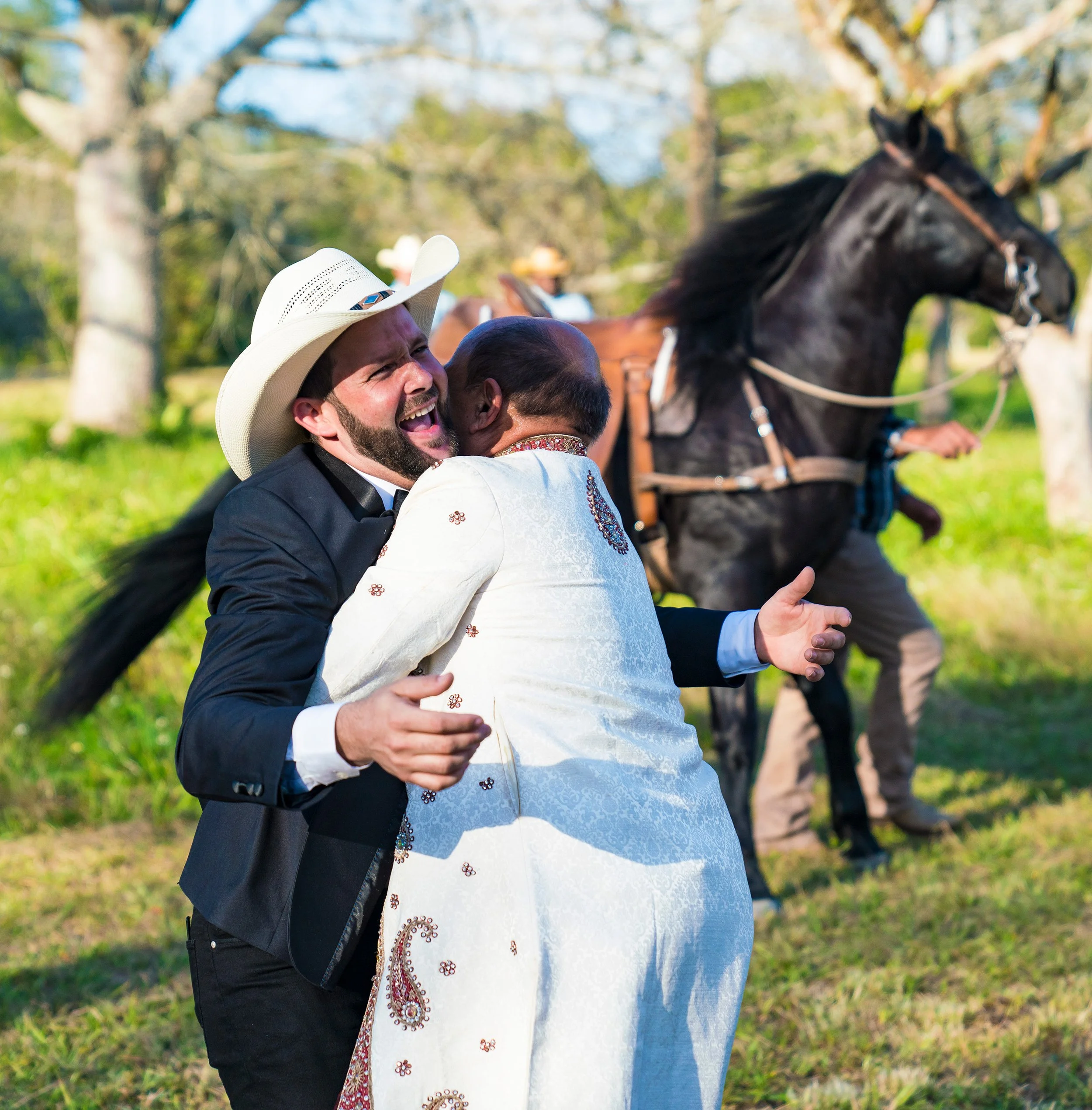 Two men embracing outdoors, one in a white cowboy hat, the other in an ornate white outfit, with a dark horse in the background.