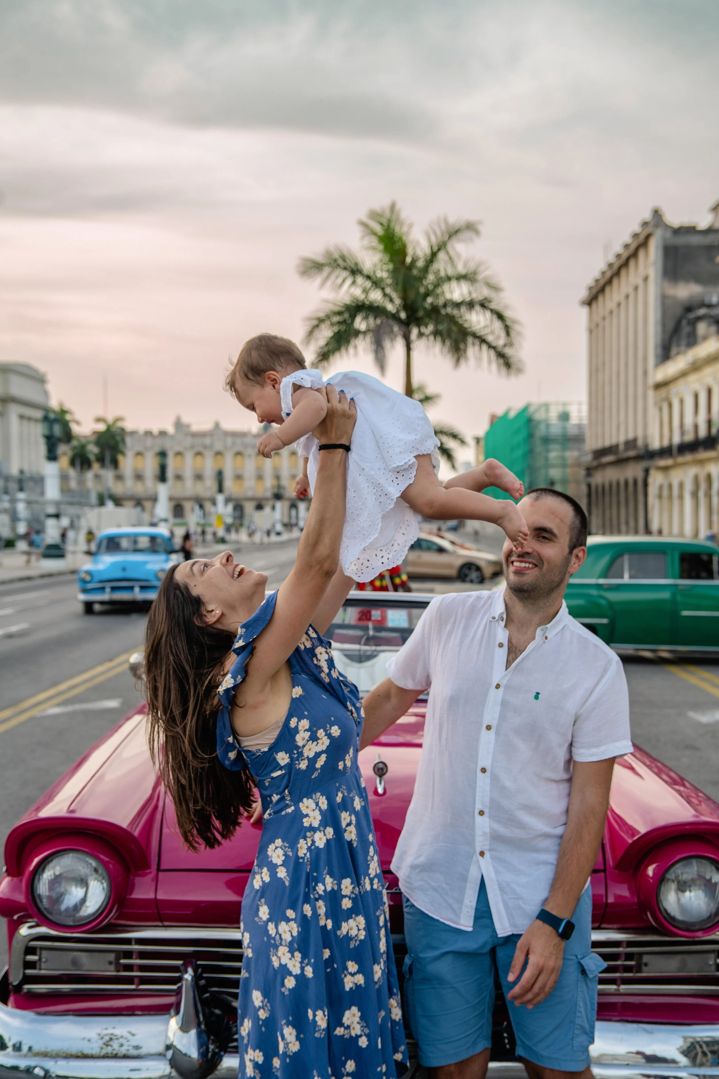 A woman and a man are smiling and holding a young girl above their heads in front of a pink vintage car, with city buildings and palm trees in the background during sunset.