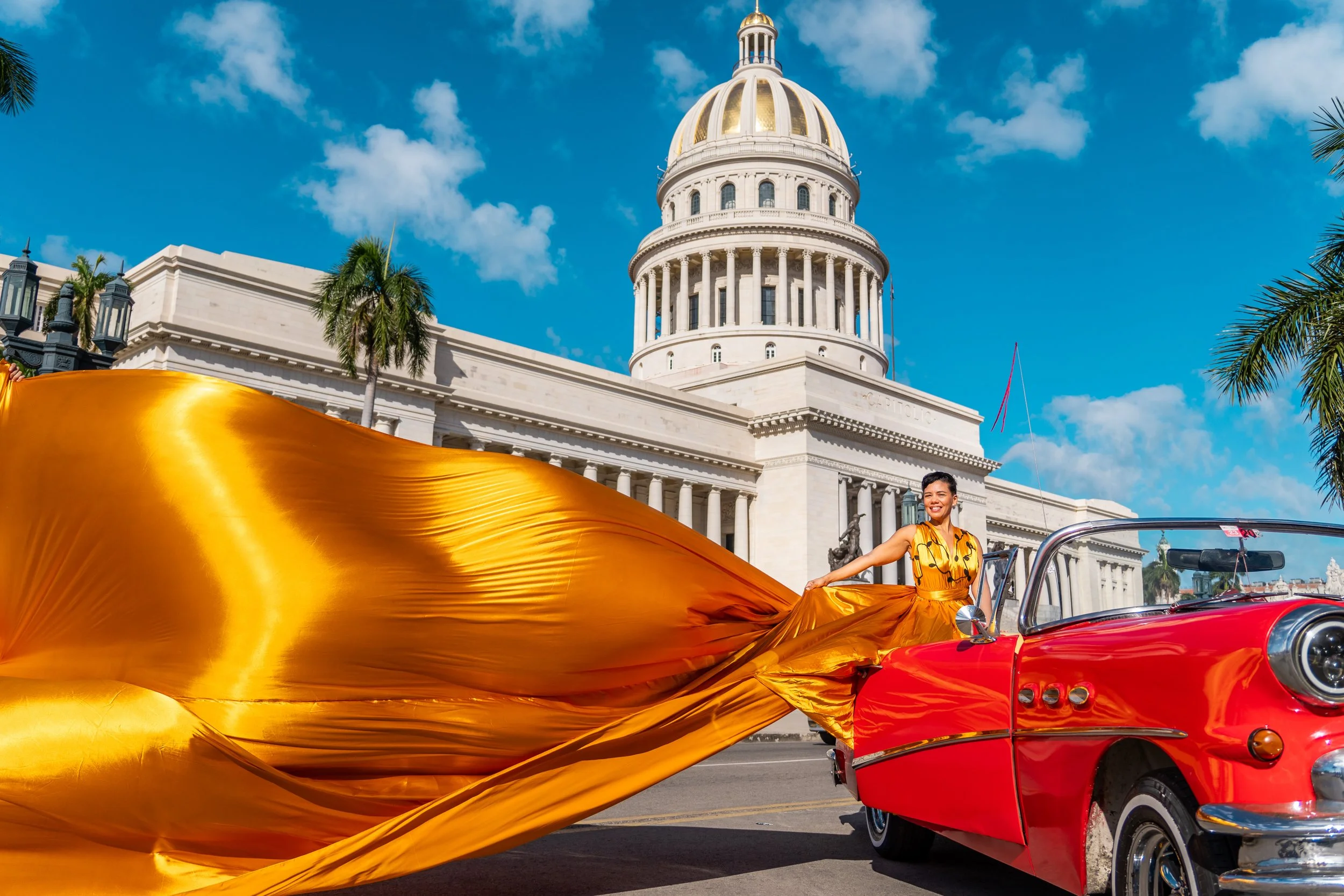 Woman in yellow dress sitting on the edge of a red convertible car with a flowing gold fabric behind her, in front of a historic white domed building with palm trees and a blue sky.