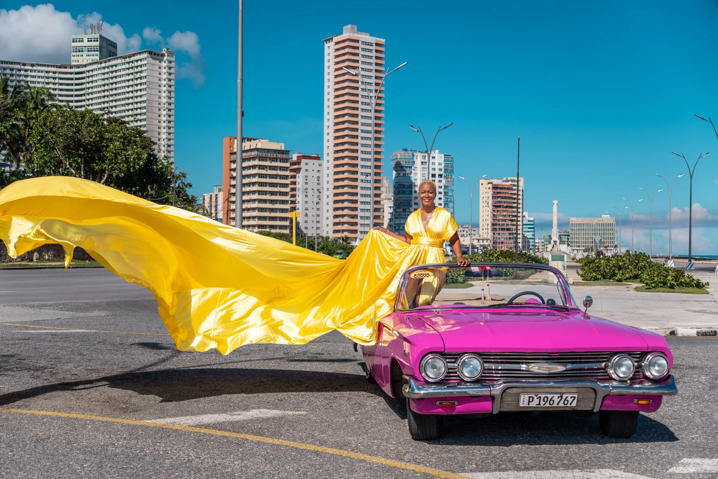 A woman in a yellow gown sitting on the pink convertible car with a yellow train flowing behind her, in an urban cityscape with tall buildings and a clear blue sky.