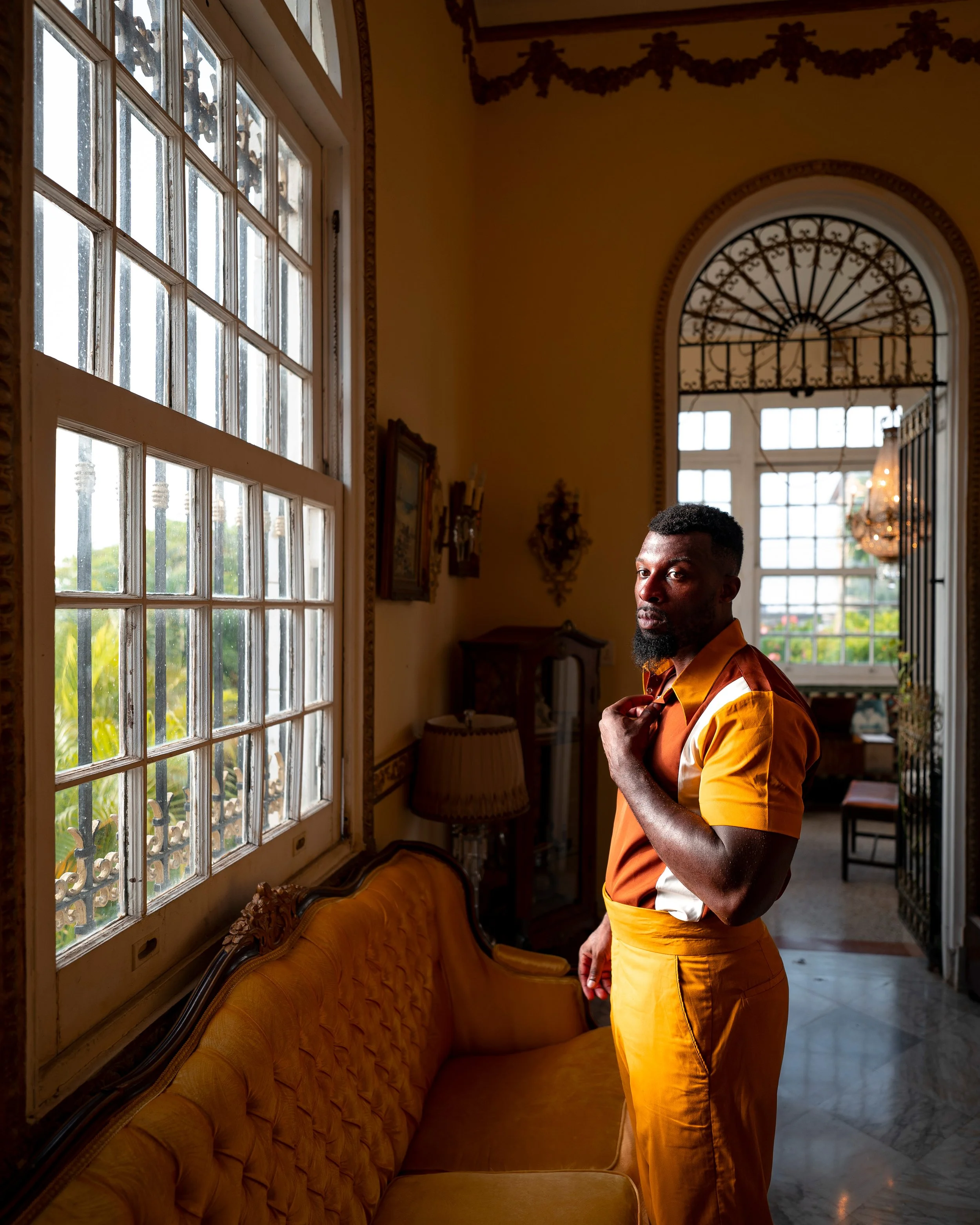 Person in vintage-inspired orange outfit standing near a window with sunlight streaming in, in a room with ornate decor and a yellow upholstered couch.