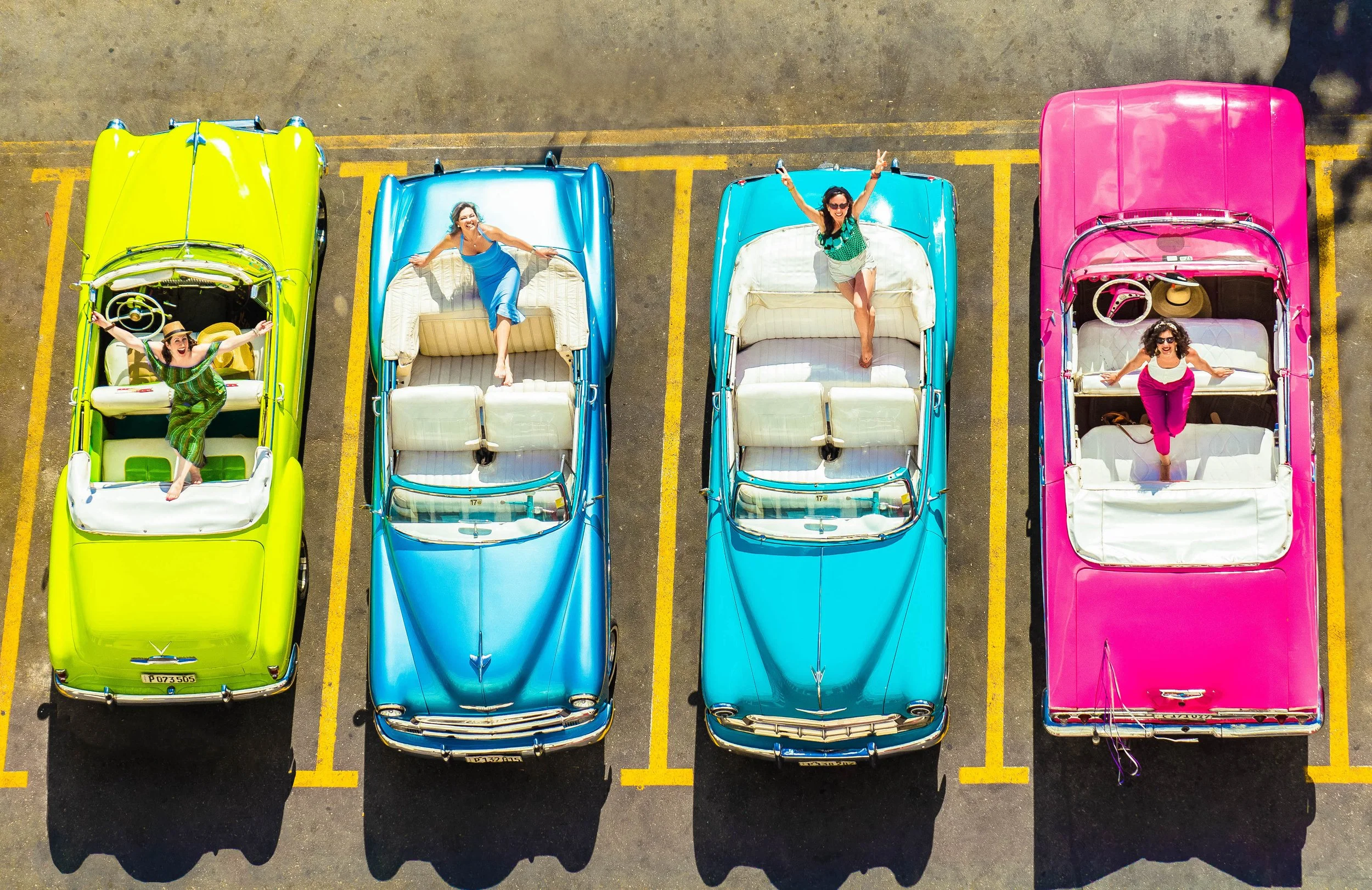 From an aerial view, four vintage convertible cars parked in a parking lot with four women standing or lying on the cars' interiors, smiling and making gestures.