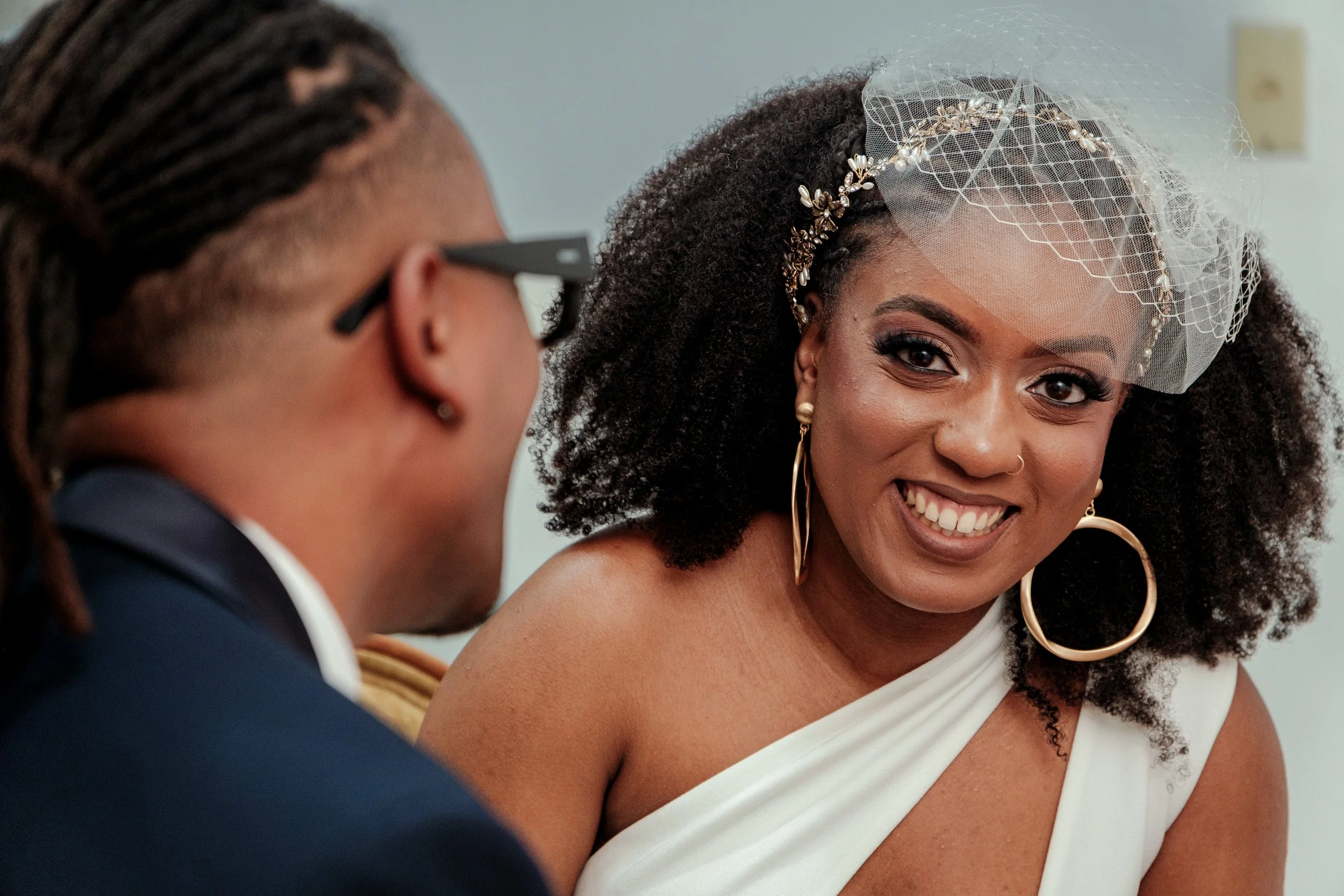 A woman wearing a wedding dress and a netted veil with a decorative headband, smiling and looking at a man with glasses and dreadlocks, who is partially visible.