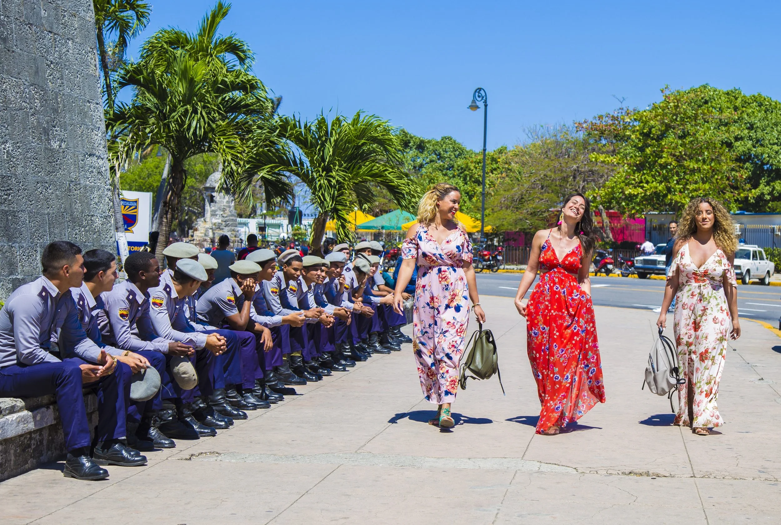 Three women in colorful dresses walking past a row of uniformed officers sitting on a low wall on a sunny day with green trees, streetlights, and parked cars in the background.