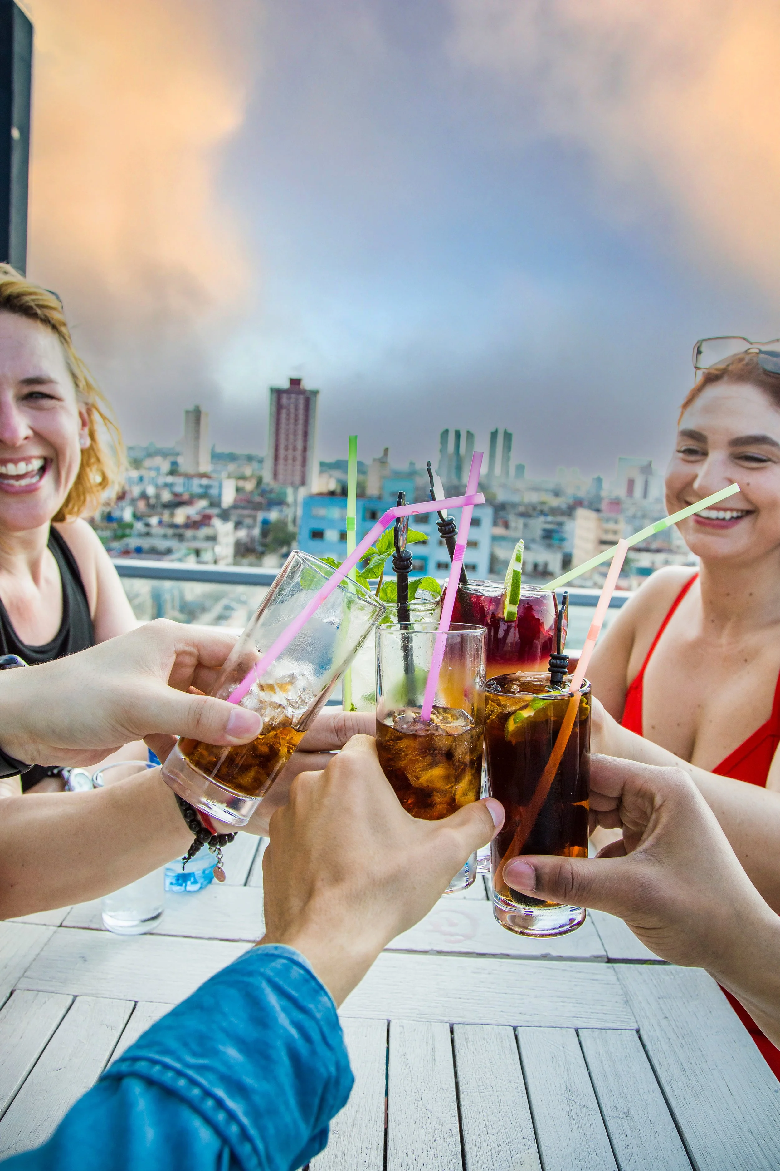 People raising glasses of mixed drinks with colorful straws on a rooftop, city skyline in the background, smiling women enjoying a celebration.